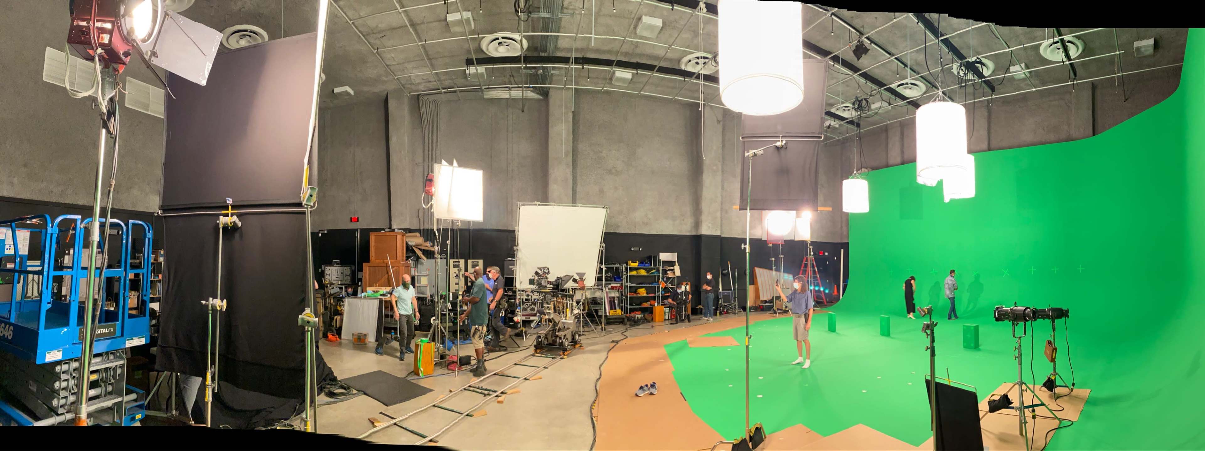 The image shows a film studio with green screens, lighting equipment, and crew members preparing for a shoot.