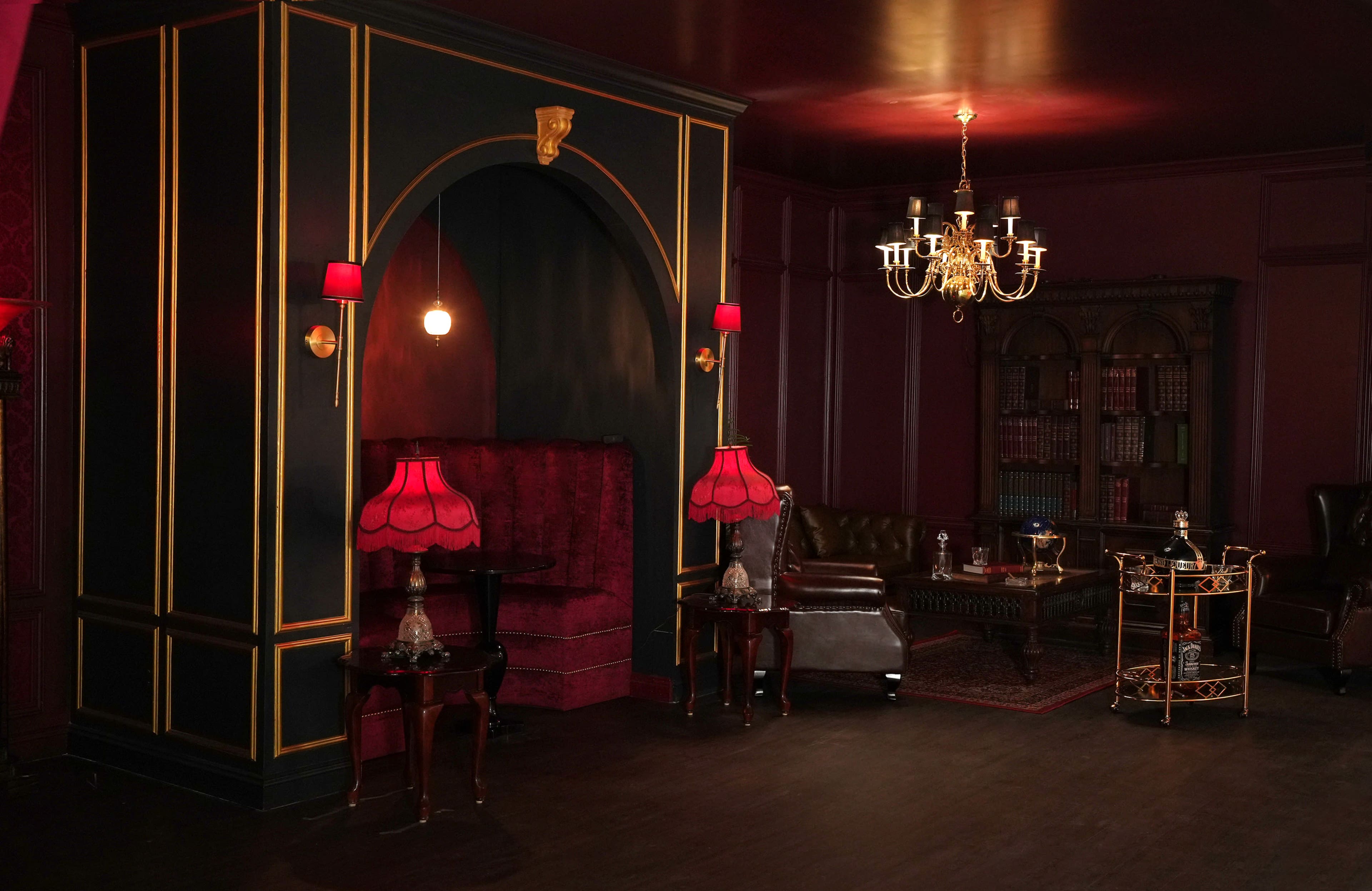 A dimly lit room with dark walls, a chandelier, two red lamps, and a mix of vintage furniture including a leather sofa and wooden tables.