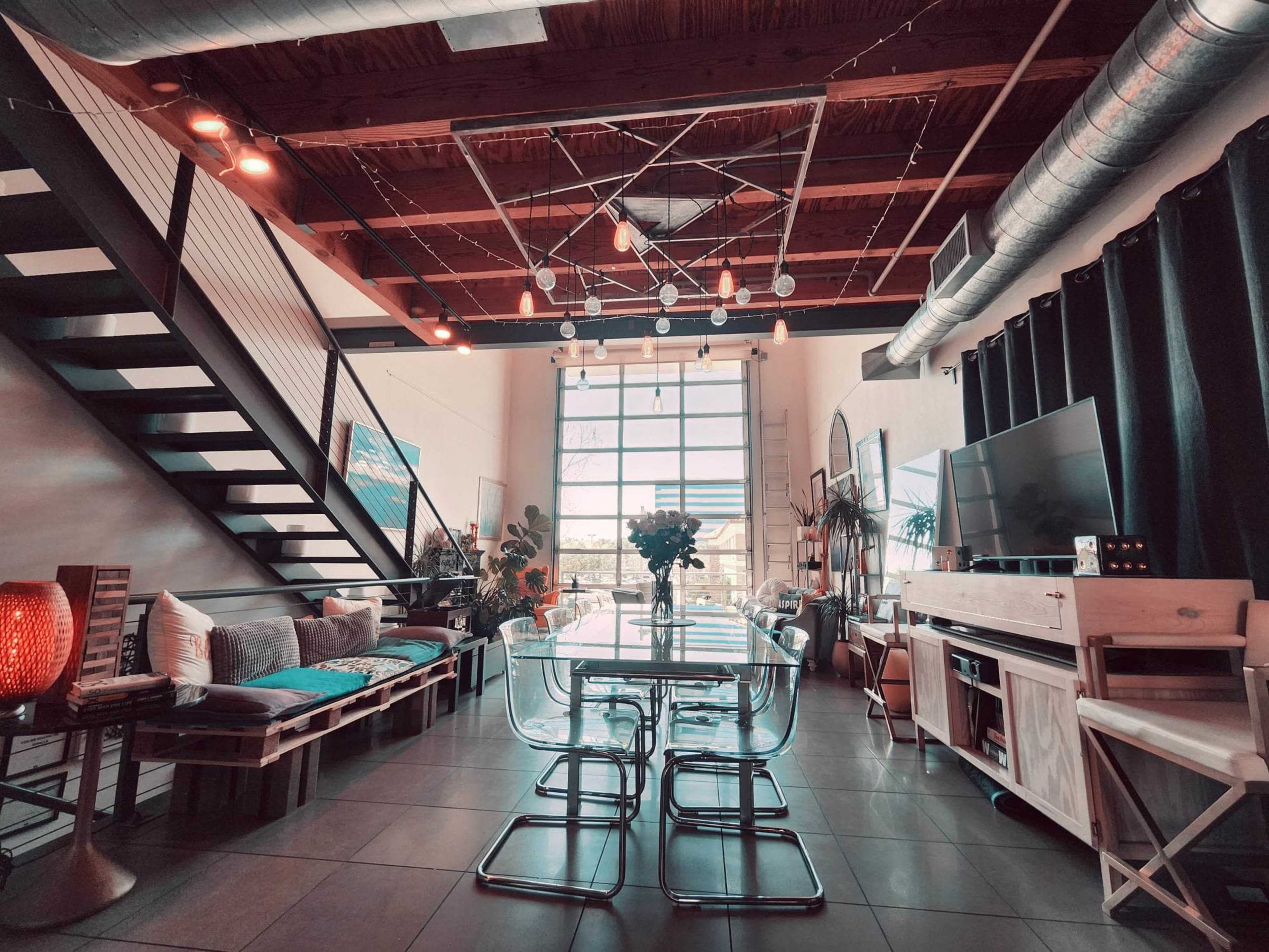 A modern loft interior features a dining area with a glass table and industrial lighting, surrounded by a staircase, seating, and decorative plants.