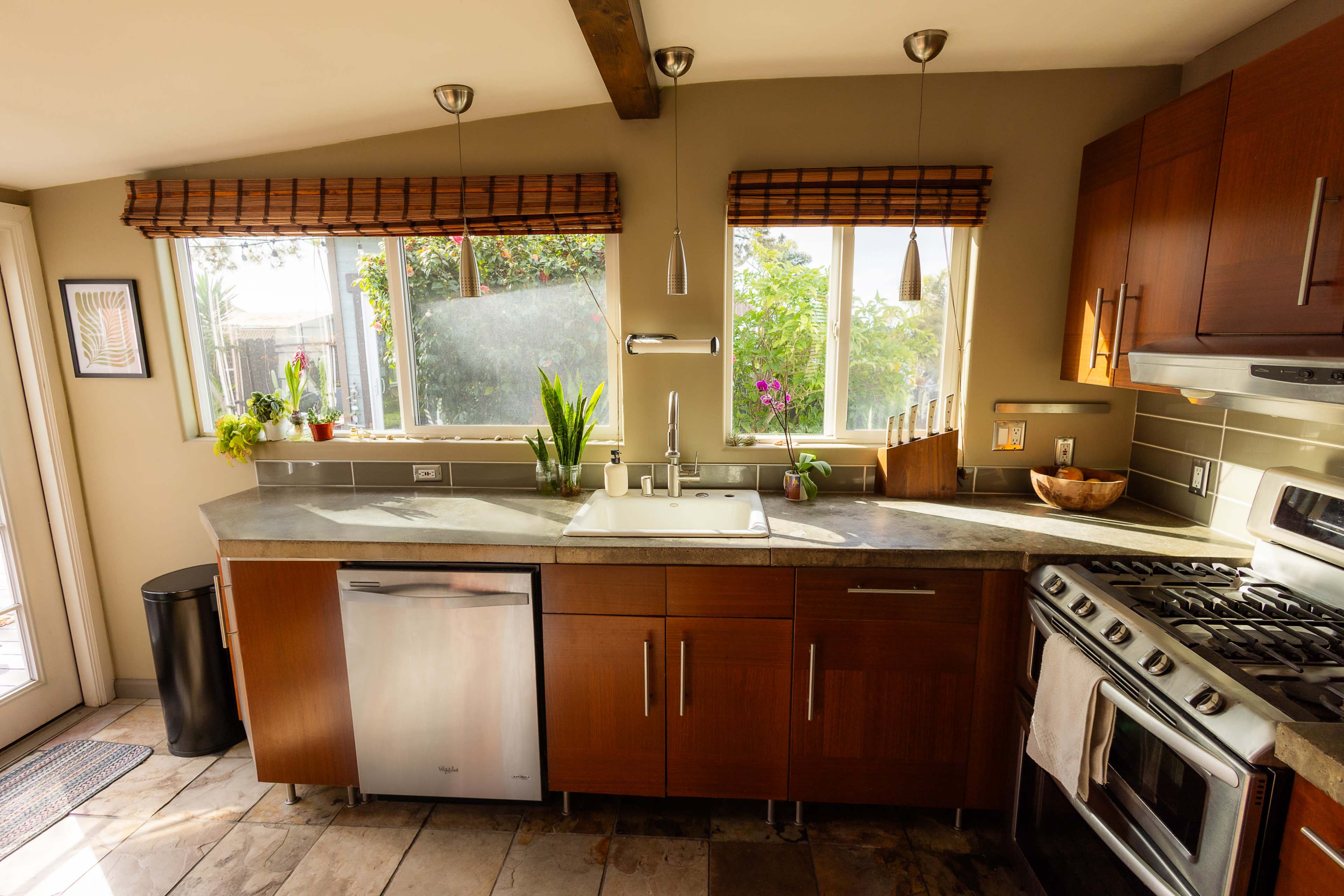 A modern kitchen with wooden cabinets, a stainless steel dishwasher, and large windows allowing natural light.