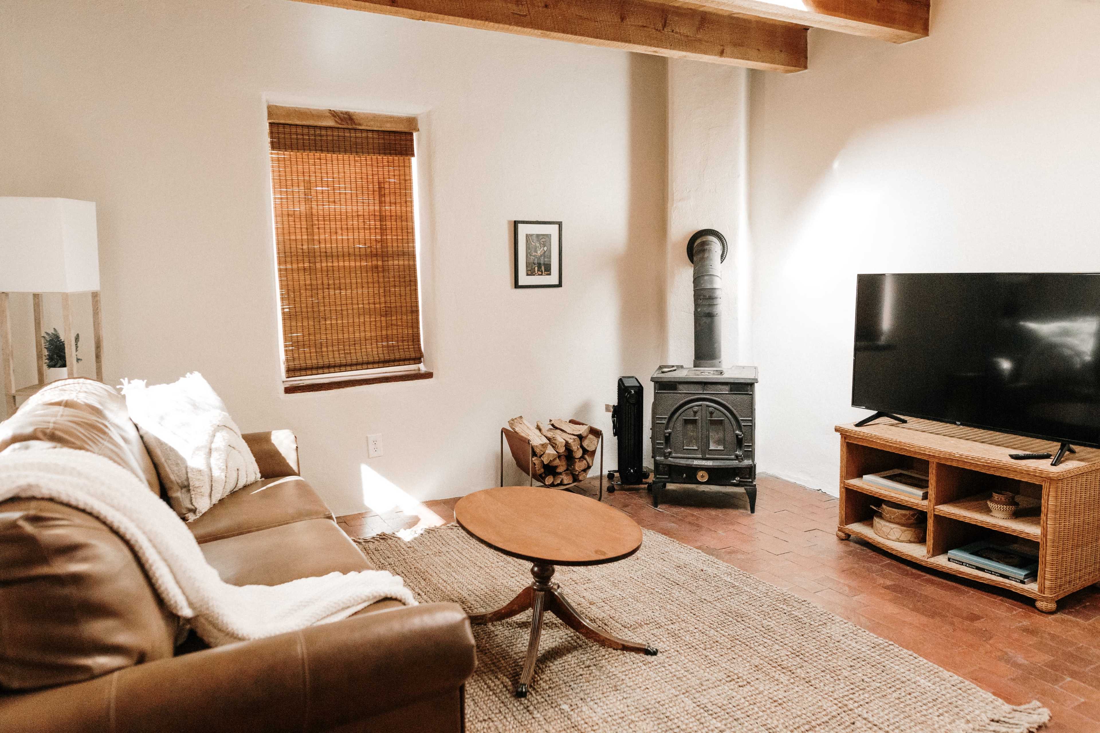 A cozy living room features a brown leather couch, a wooden coffee table, a television on a stand, and a wood-burning stove beside stacked firewood.