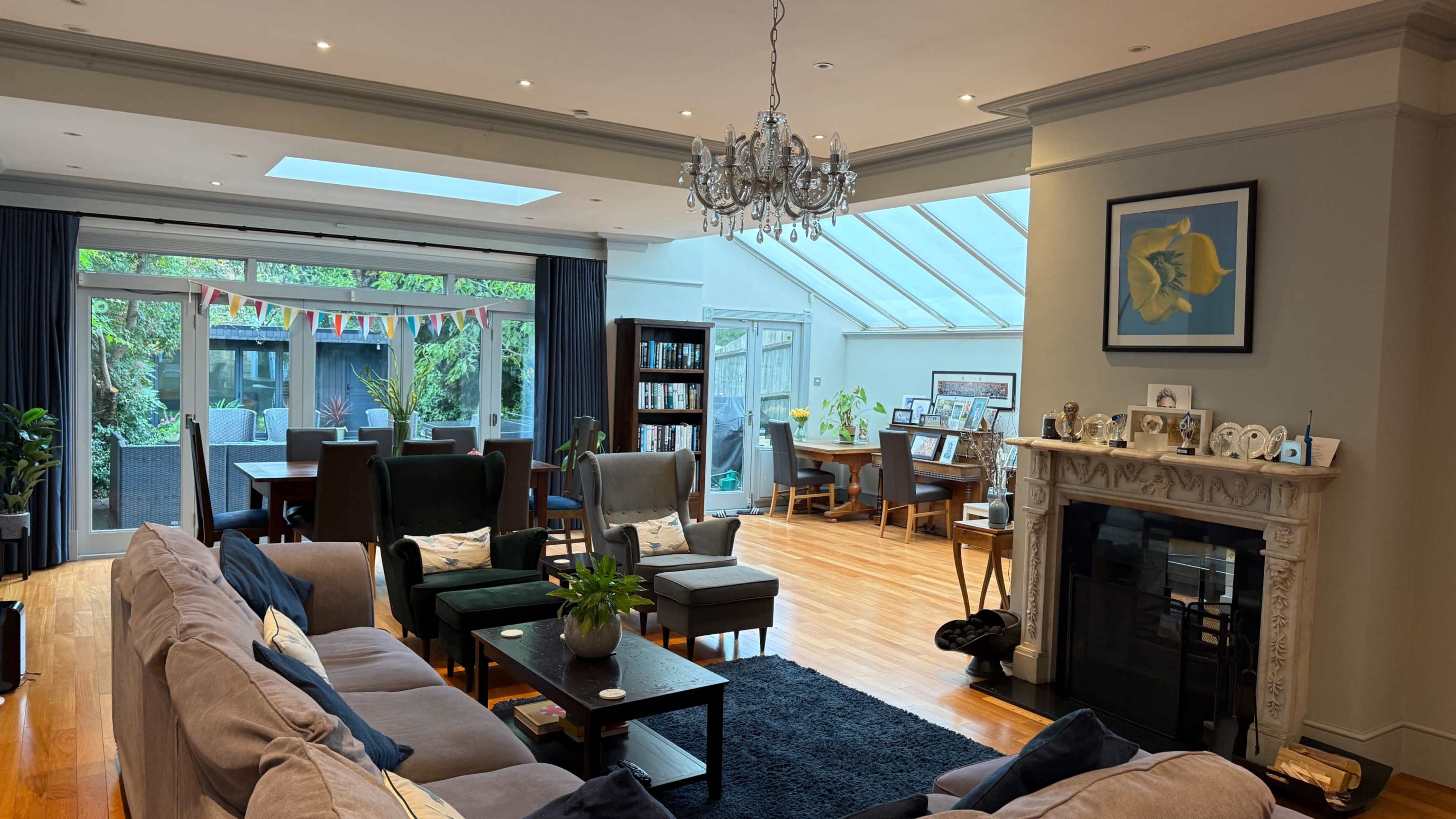 The image shows a spacious living room with modern furnishings, a fireplace, a large skylight, and a dining area visible through glass doors.