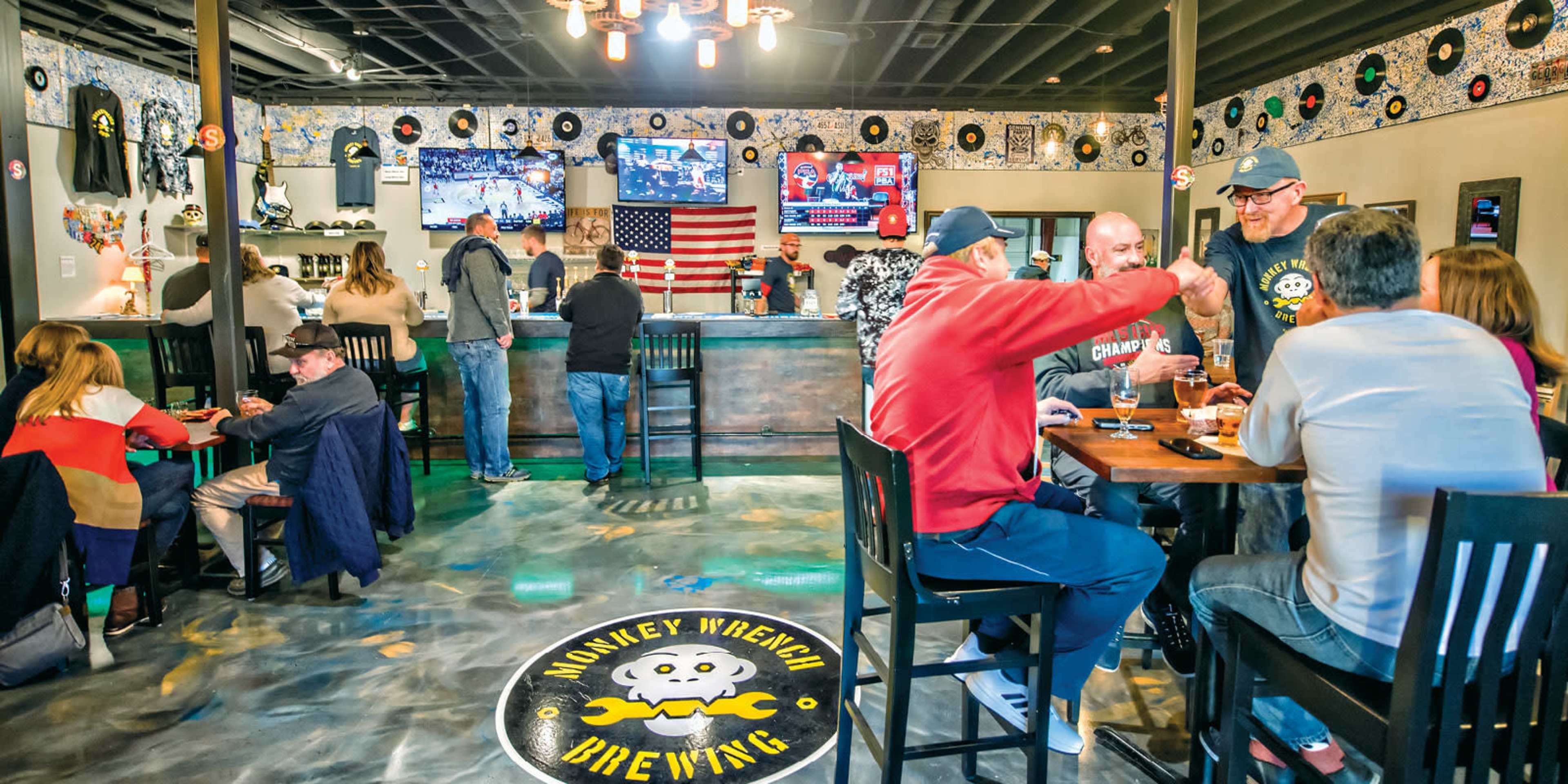 A brewery interior features patrons socializing at tables and a bar, with televisions displaying sports and a large American flag on the wall.