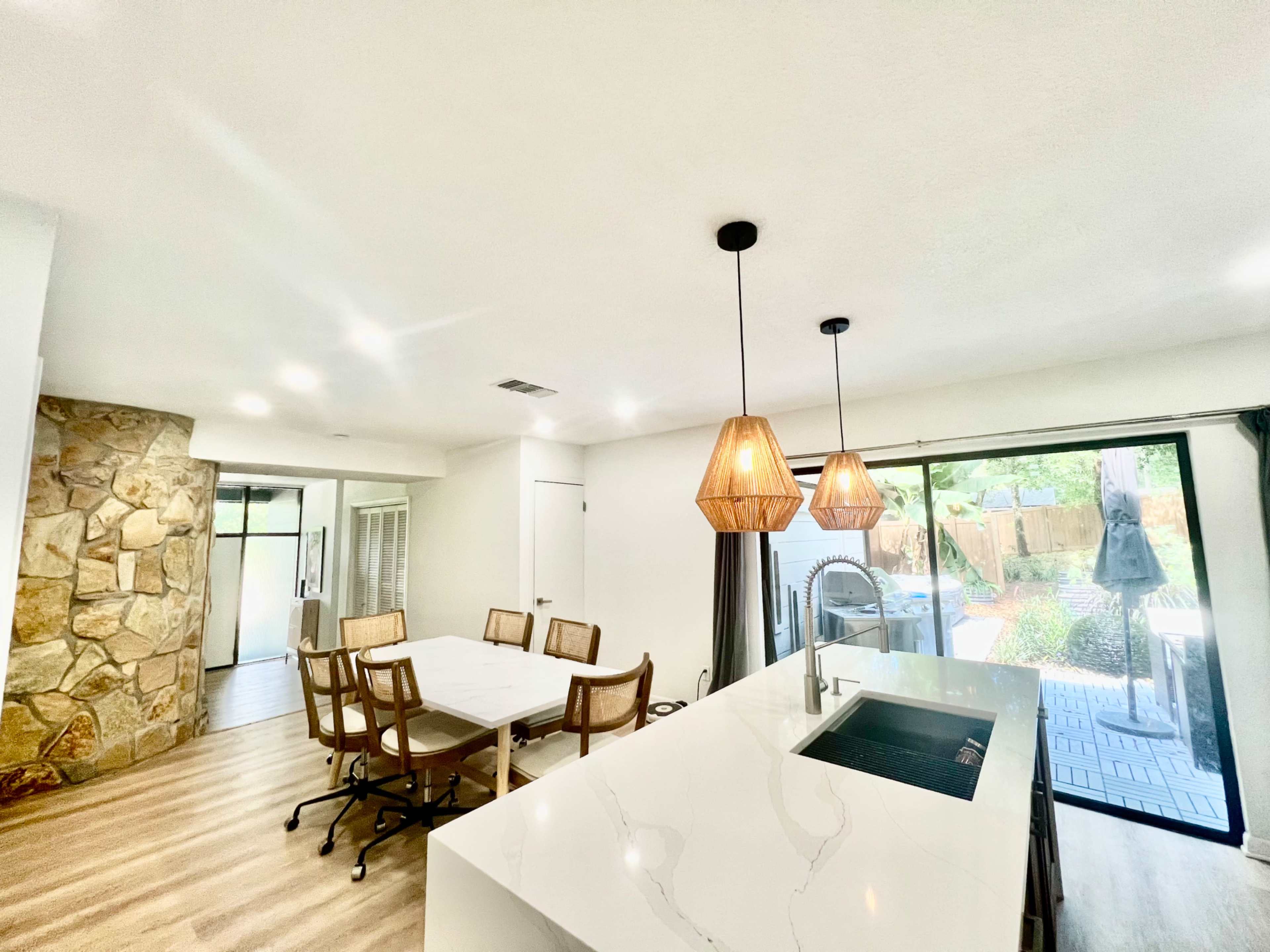 The image shows a modern interior space featuring a dining area with a large table, a kitchenette with a marble countertop, and large sliding glass doors that open to an outdoor patio surrounded by landscaping.