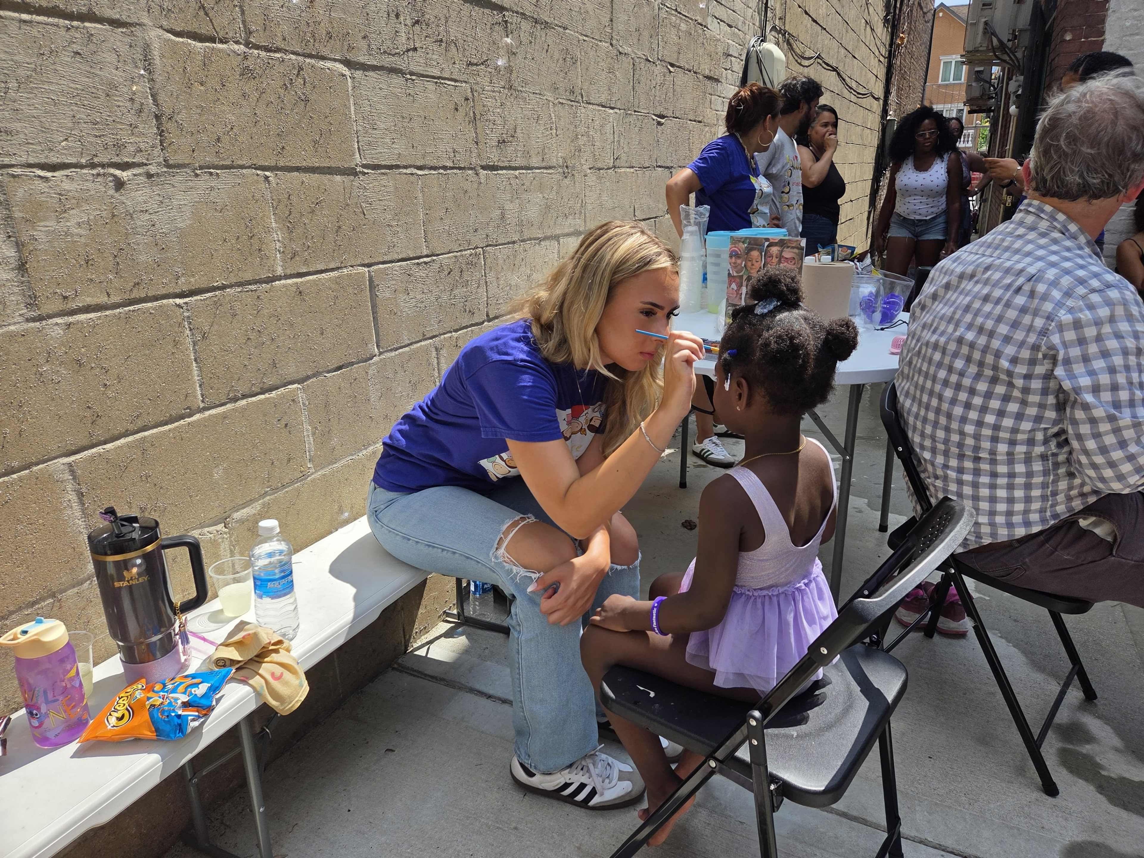 A woman is applying face paint on a young girl sitting in a chair in an outdoor setting with people and tables in the background.