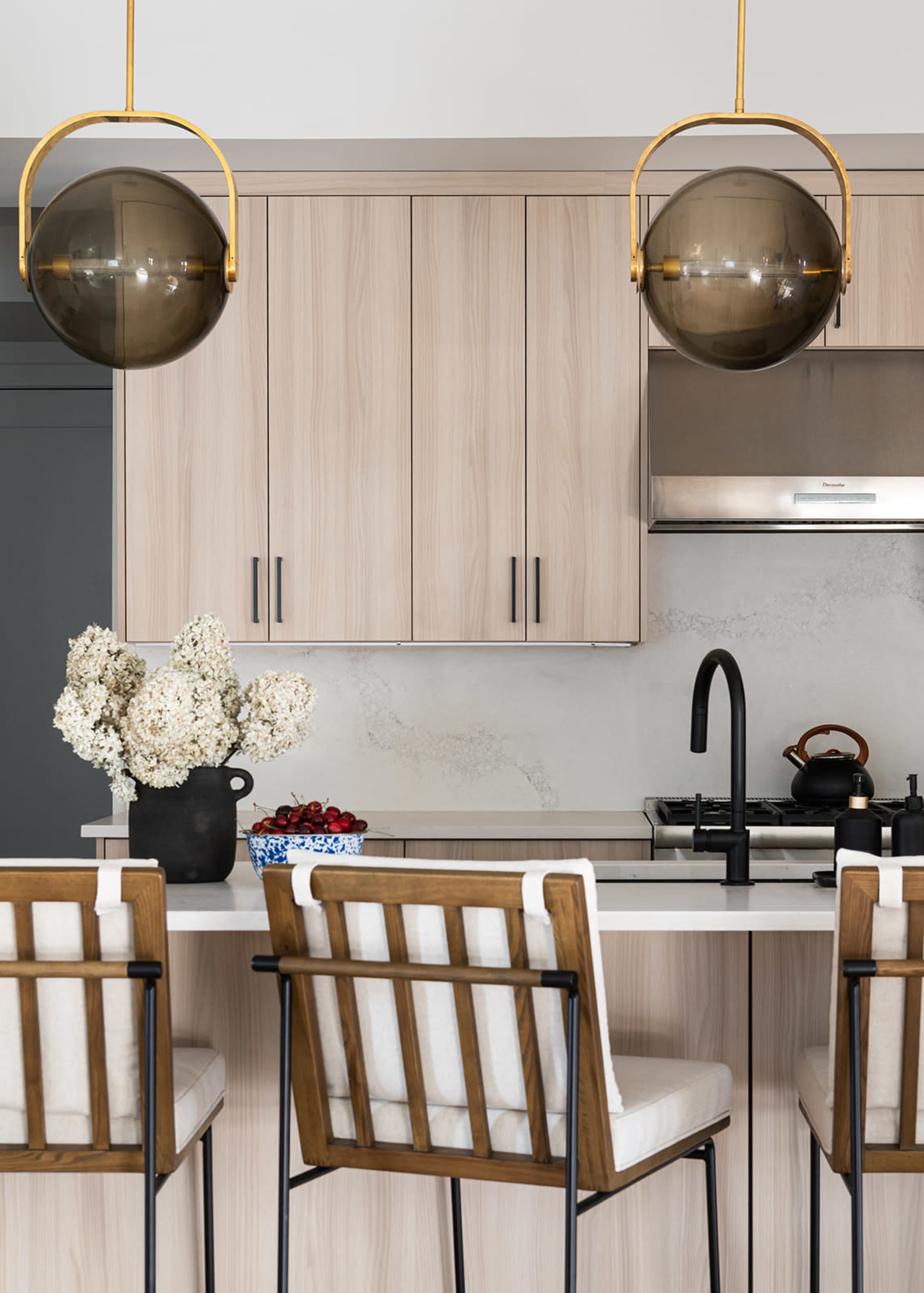 The image shows a modern kitchen with light wood cabinetry, metallic accents, and two pendant lights hanging above a countertop with bar seating.
