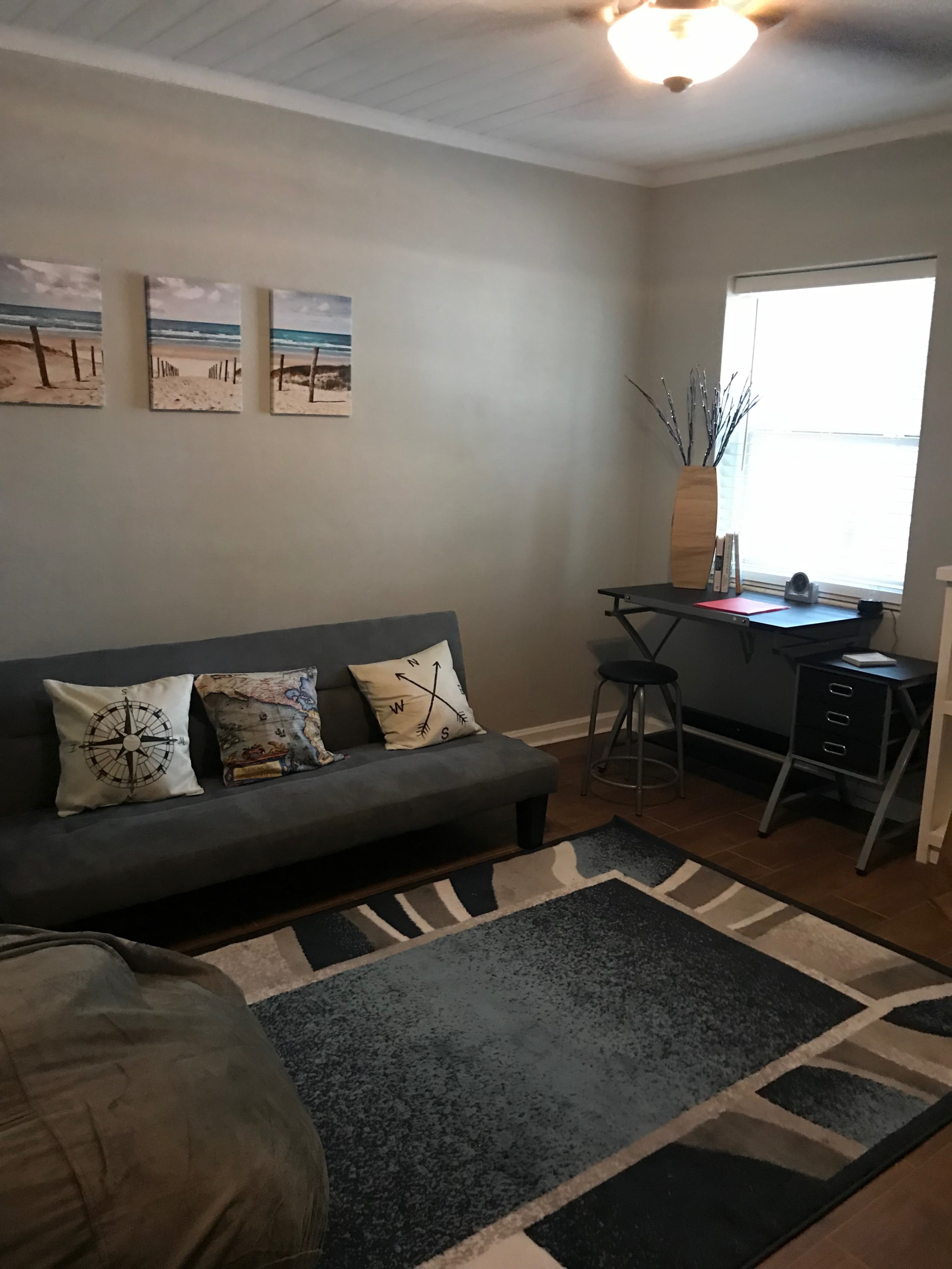 A small, well-lit living area featuring a grey futon, decorative pillows, a window with natural light, and a desk with a chair.