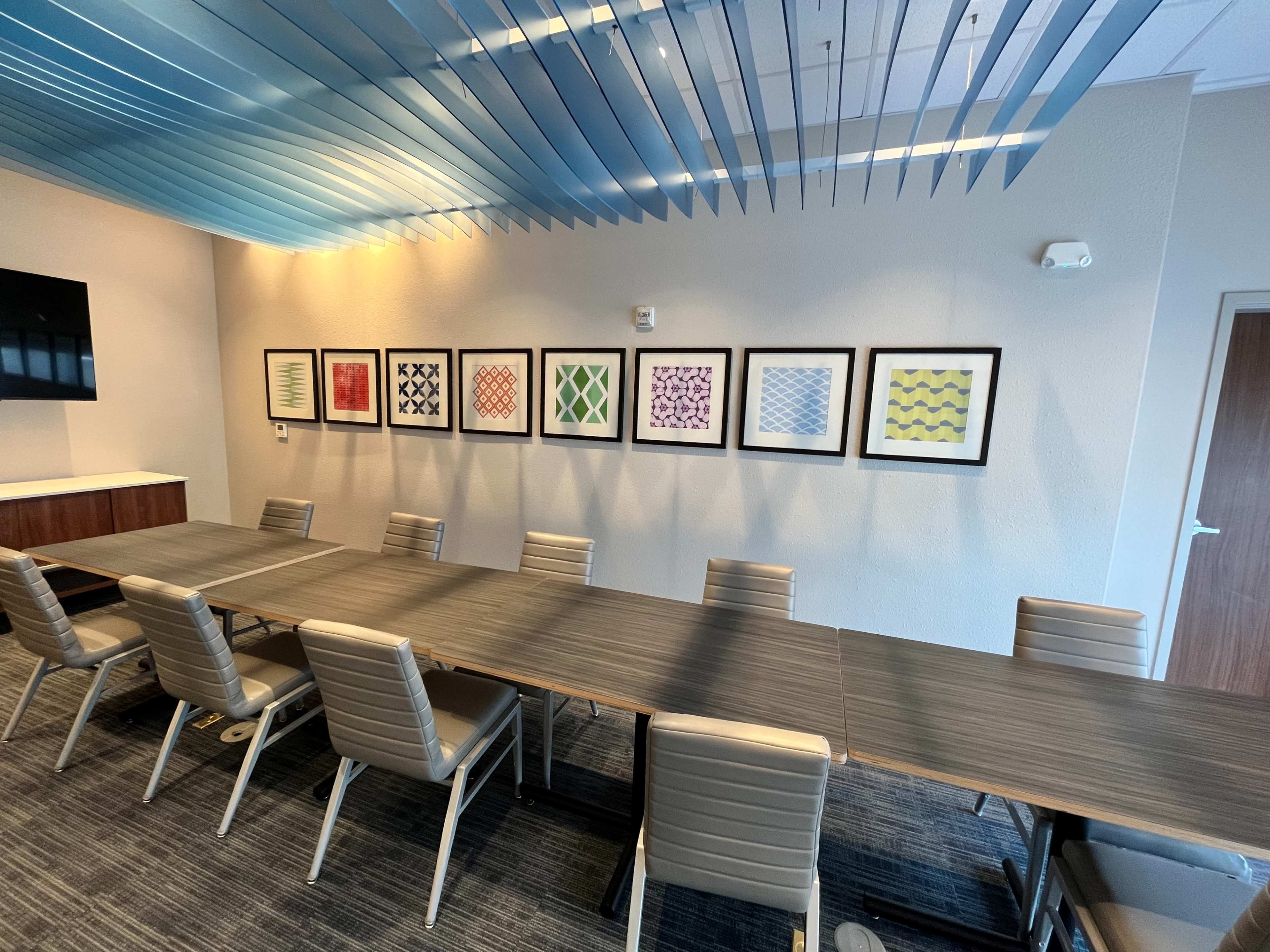 A long conference table with several chairs is set up in a meeting room, featuring blue overhead decorations and a wall adorned with framed abstract artwork.