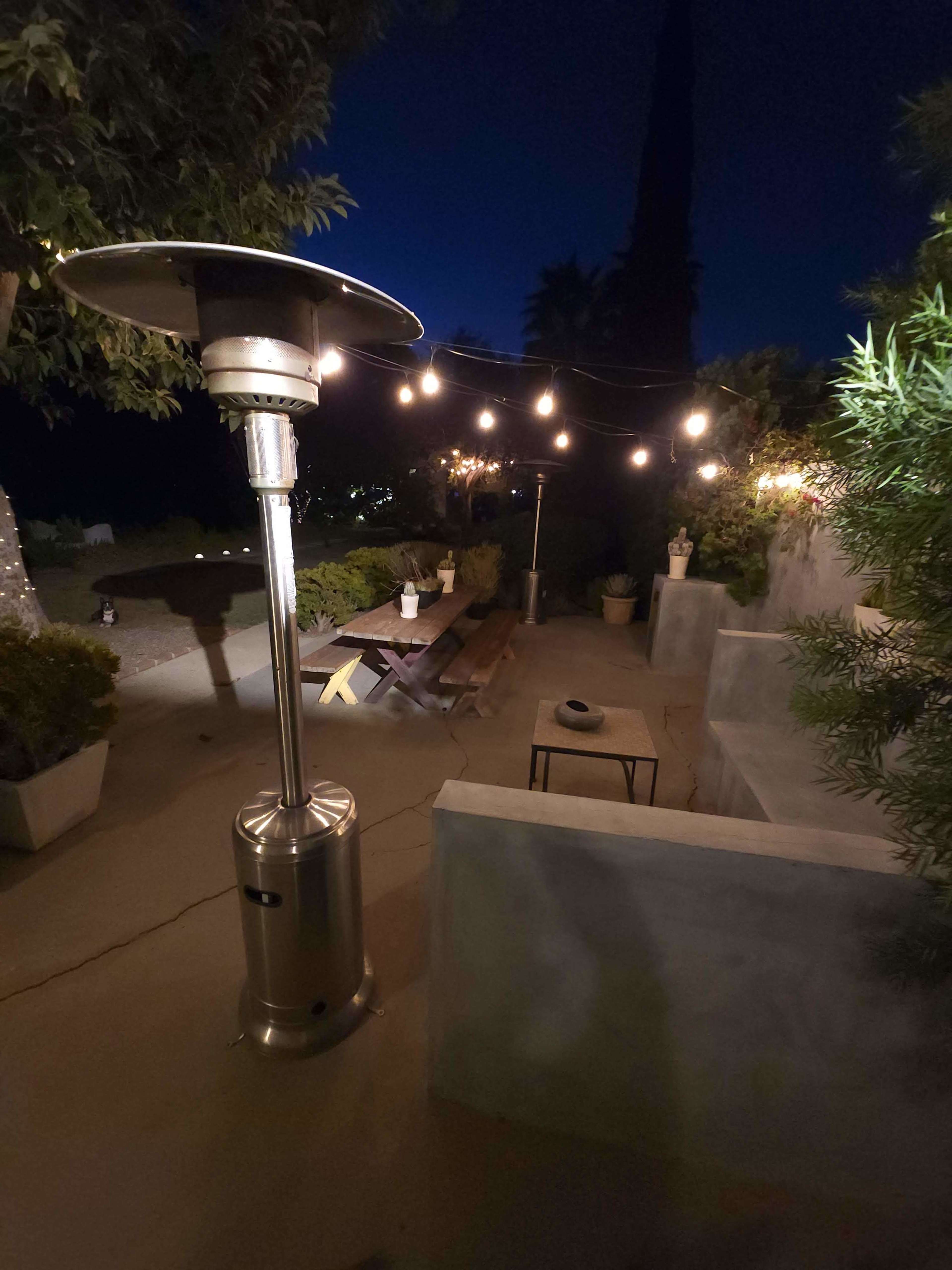 A patio area is illuminated by string lights, featuring a tabletop heater, a wooden bench, and potted plants, under a night sky.