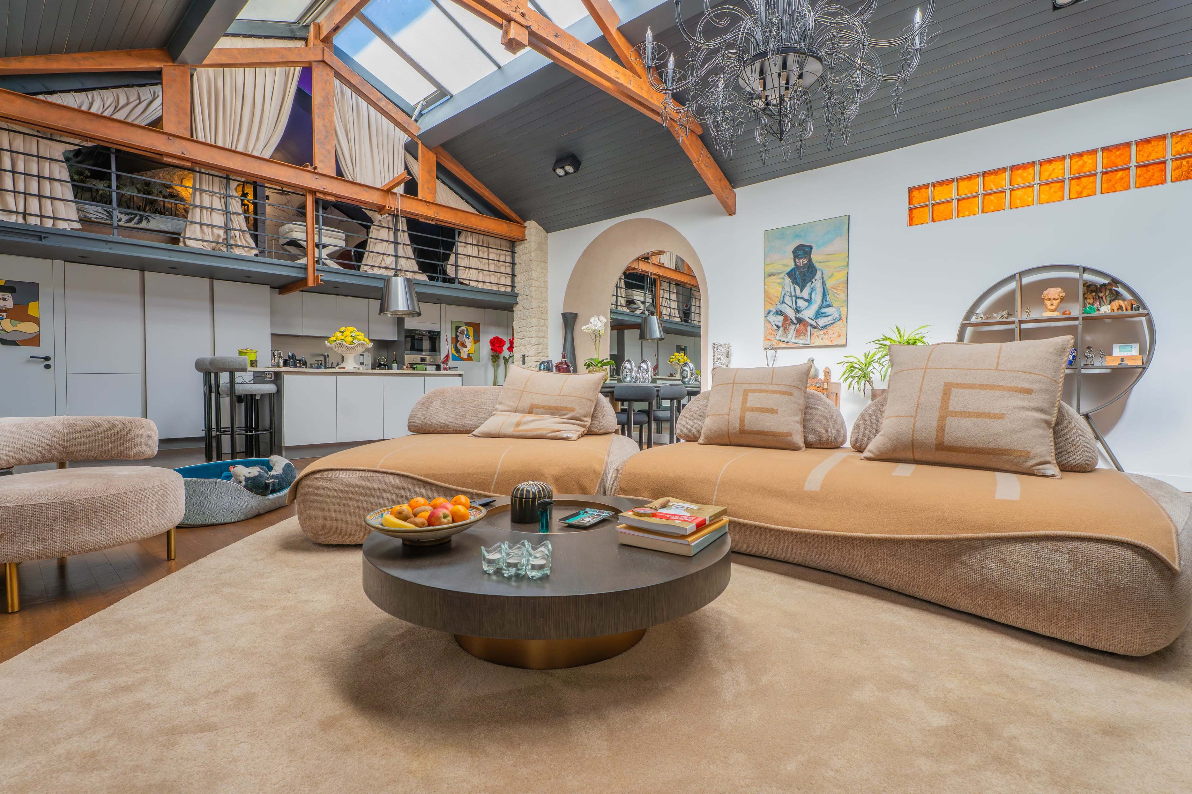 The image shows a modern living room with a large sectional sofa, a circular coffee table, and a kitchen area in the background, illuminated by natural light from a skylight.