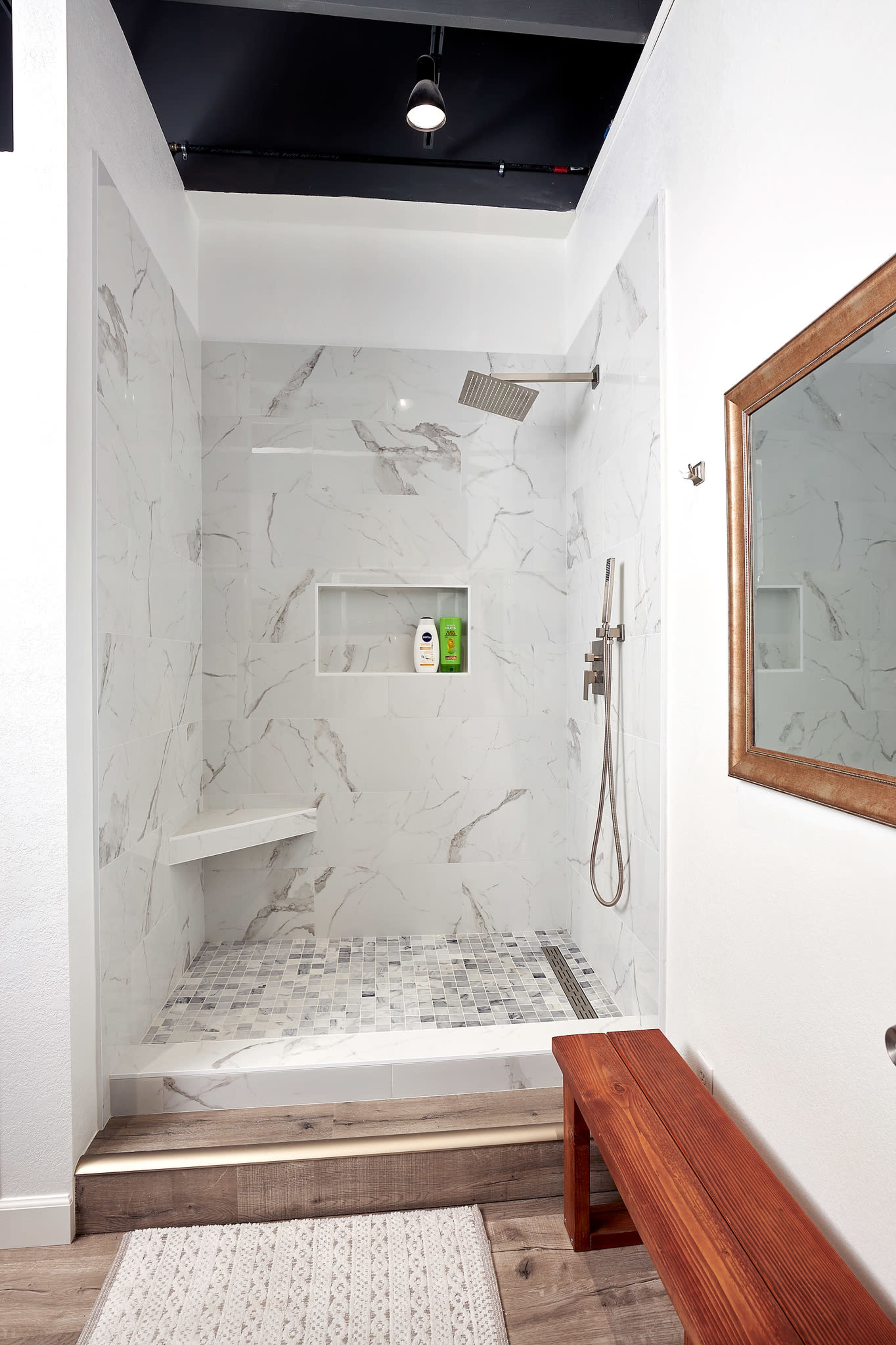 The image shows a modern shower stall with white marble walls, a glass partition, a built-in shelf for toiletries, and a wooden bench outside.