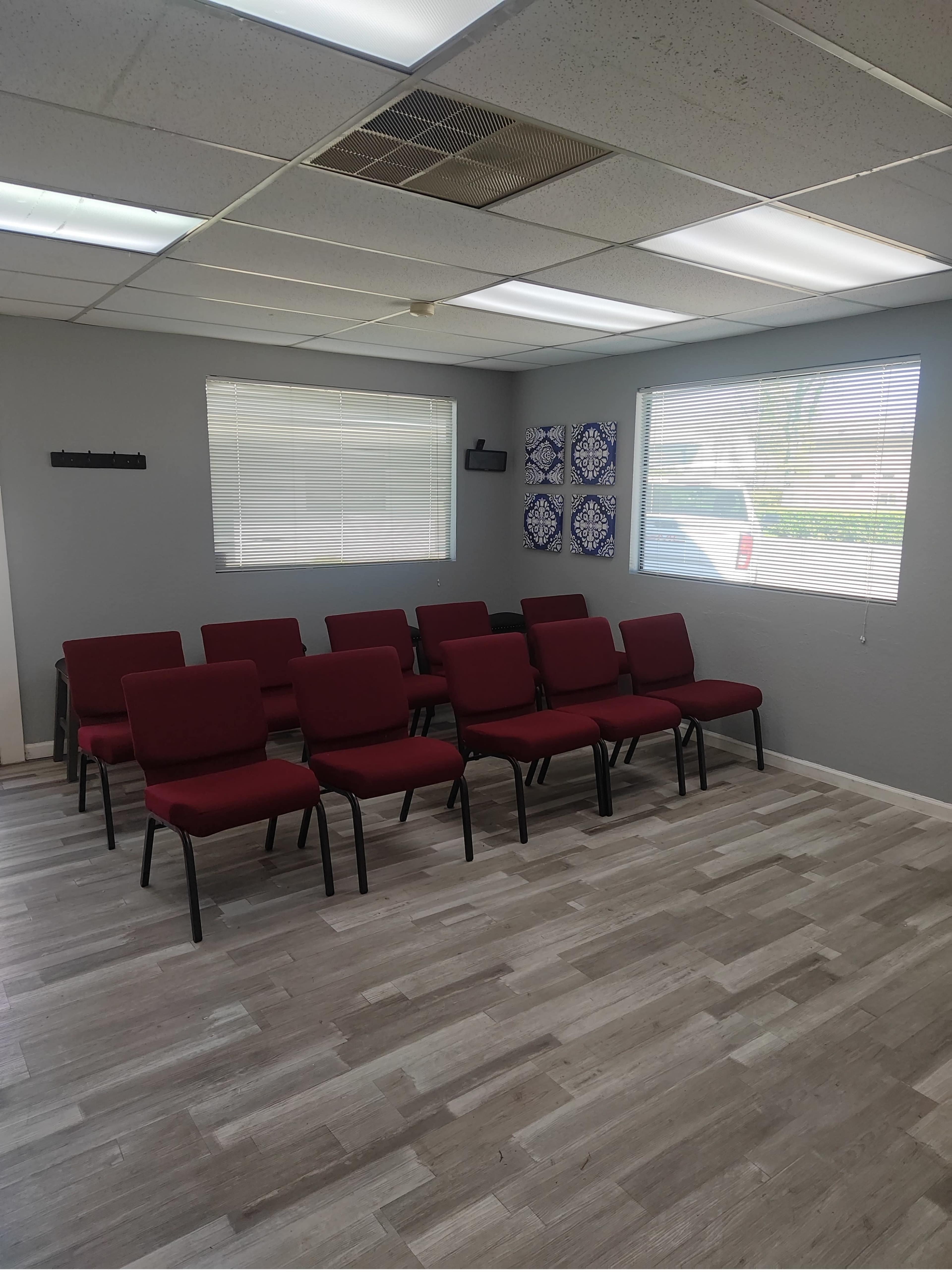 The image shows a waiting room with several red chairs arranged in neat rows, illuminated by natural light from large windows.