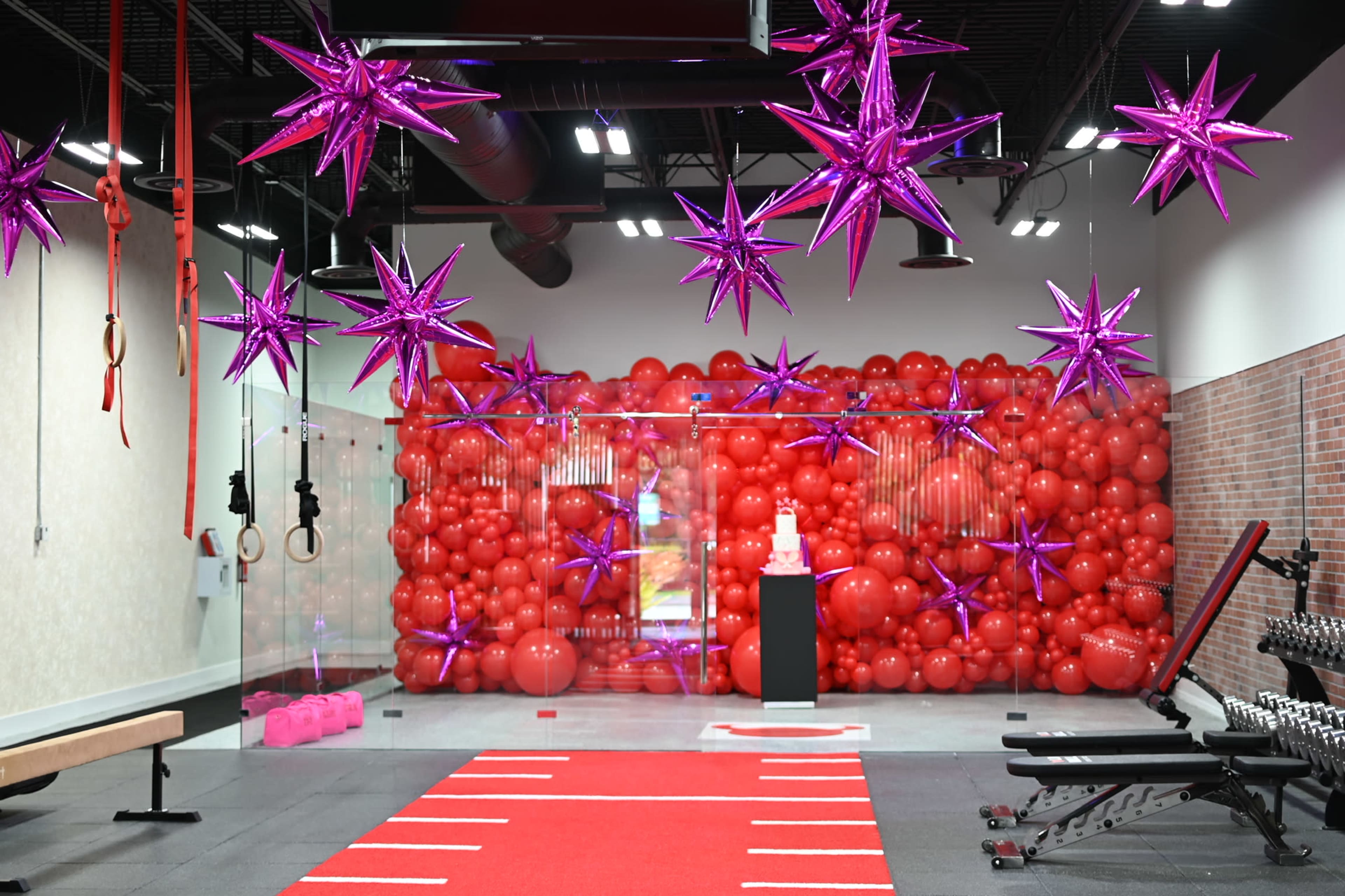A gym interior decorated with a backdrop of red balloons and purple star-shaped ornaments, featuring exercise equipment and a central cake display on a pedestal.