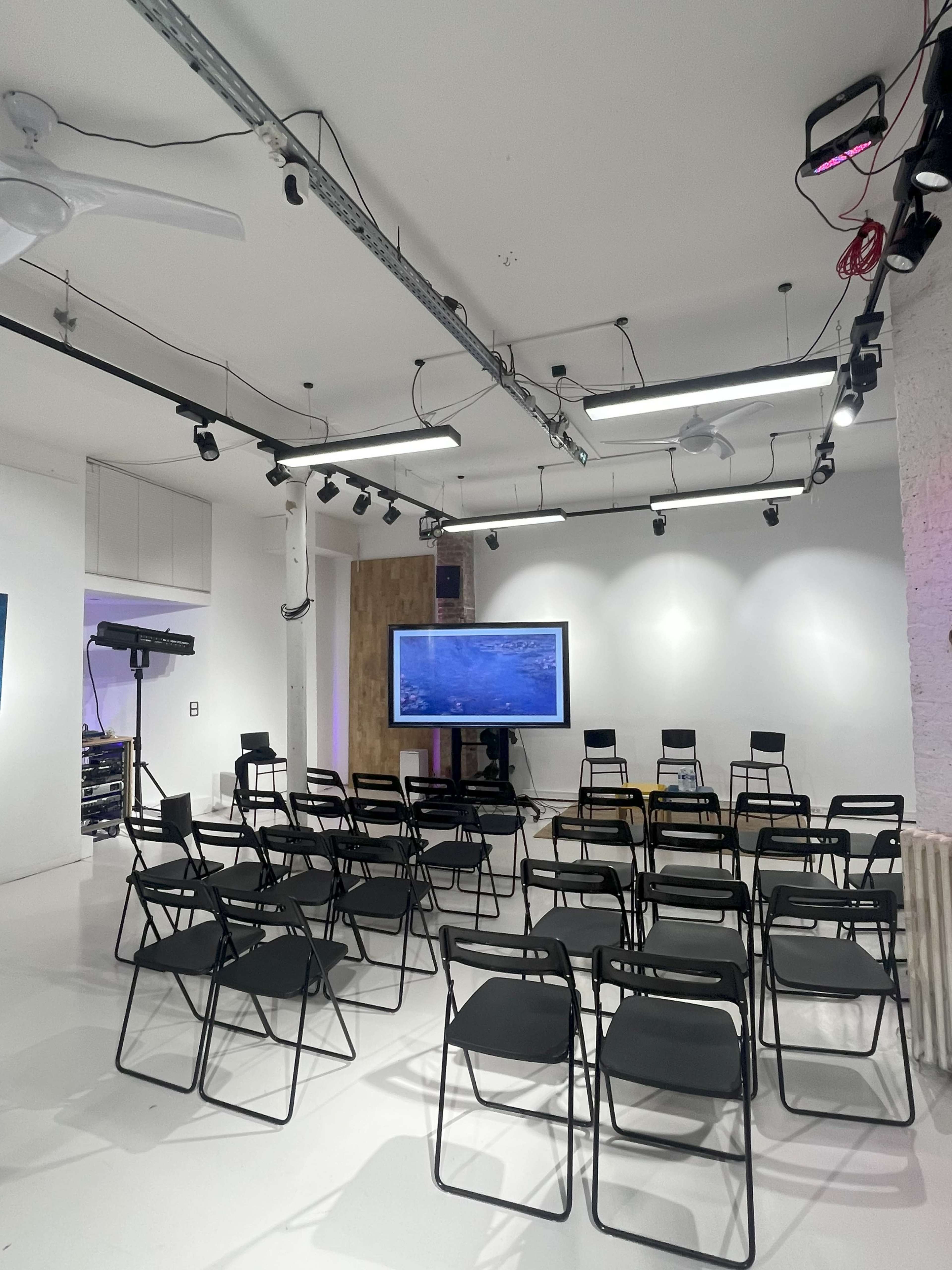 The image shows a modern, empty event space with black folding chairs arranged in rows facing a large screen.