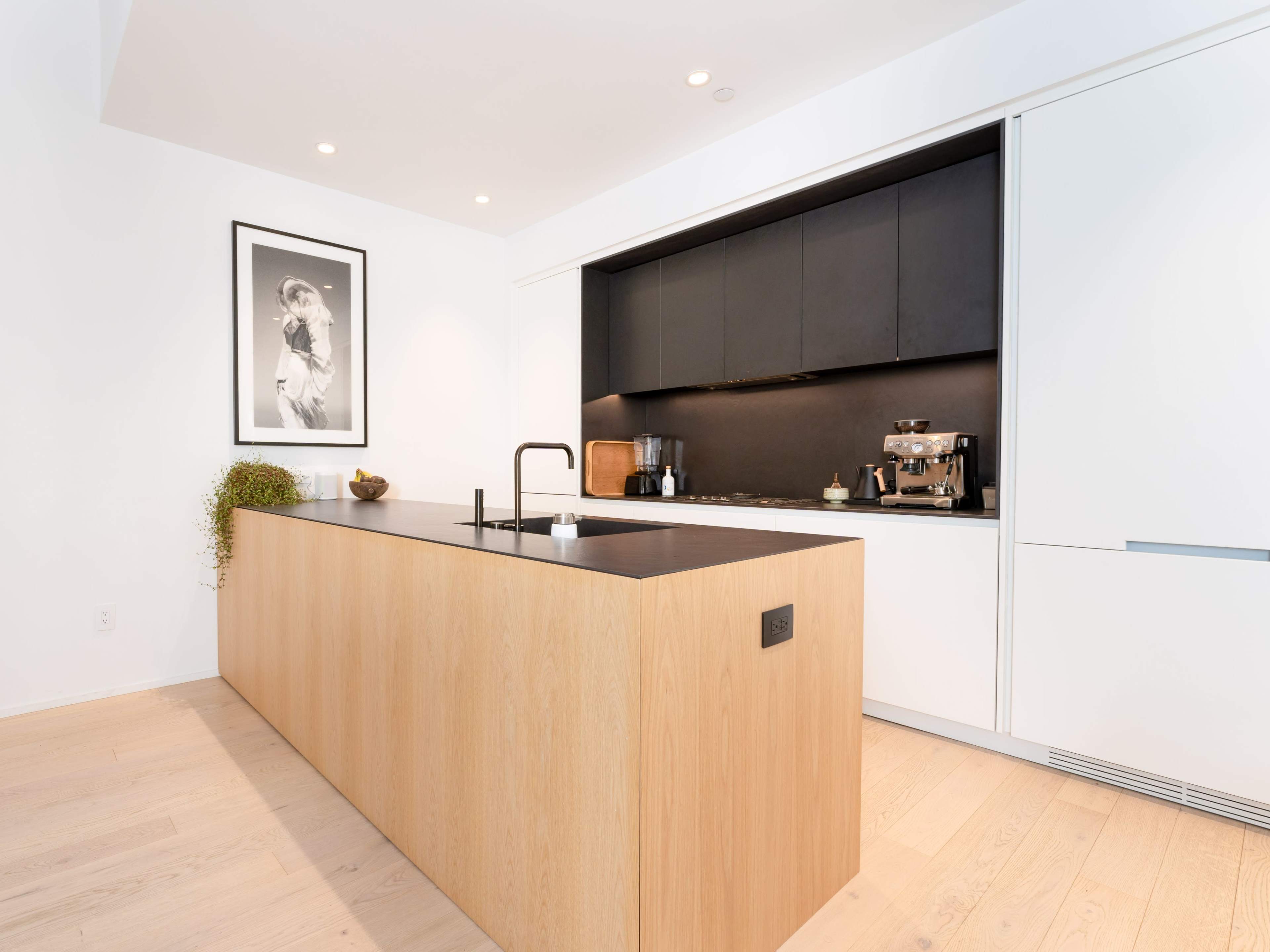 The image shows a modern kitchen featuring a large wooden island, sleek black cabinetry, and framed artwork on the wall.