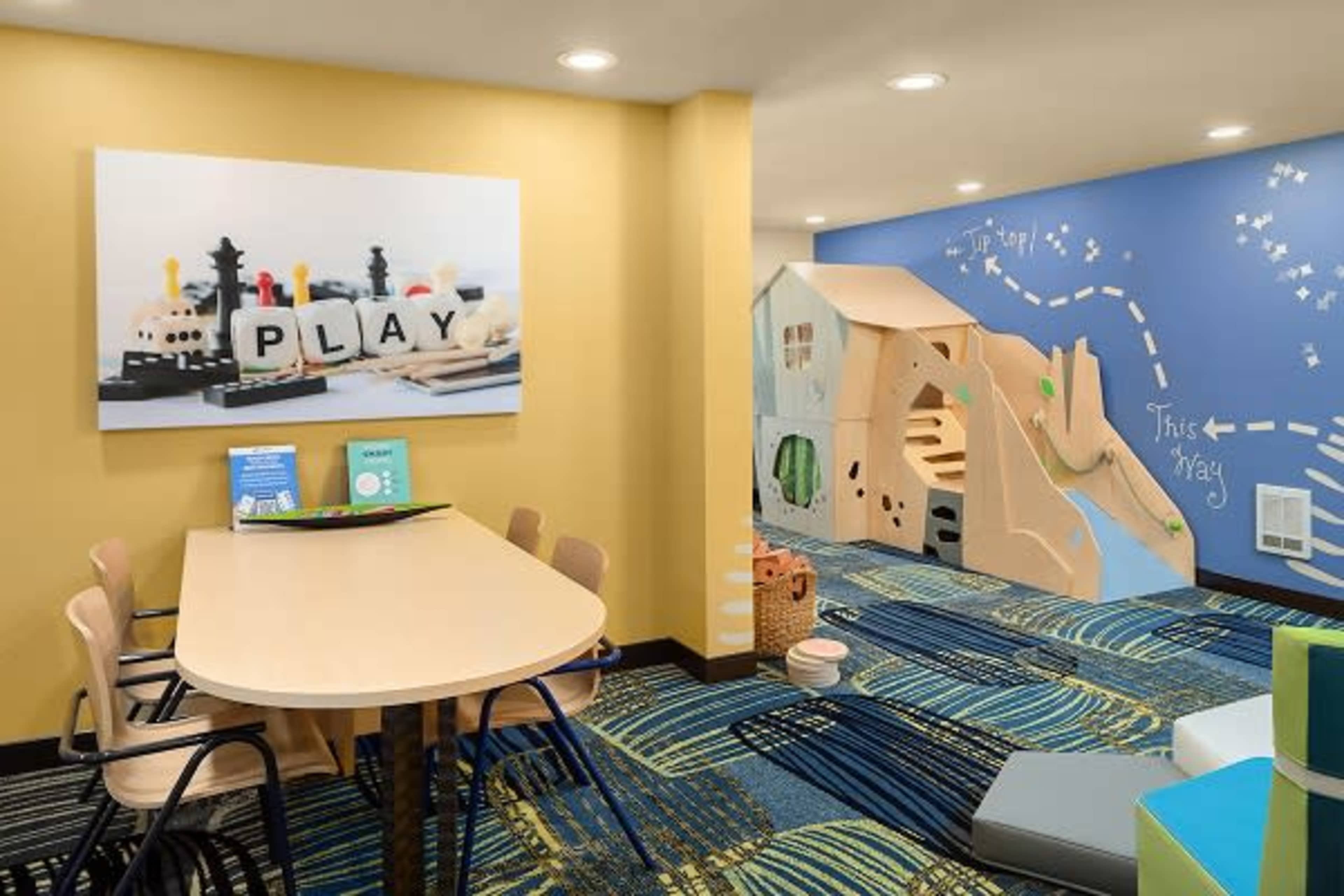 A colorful children's play area featuring a table with wooden toys and a playhouse structure against a wall with a blue mural and directional arrows.