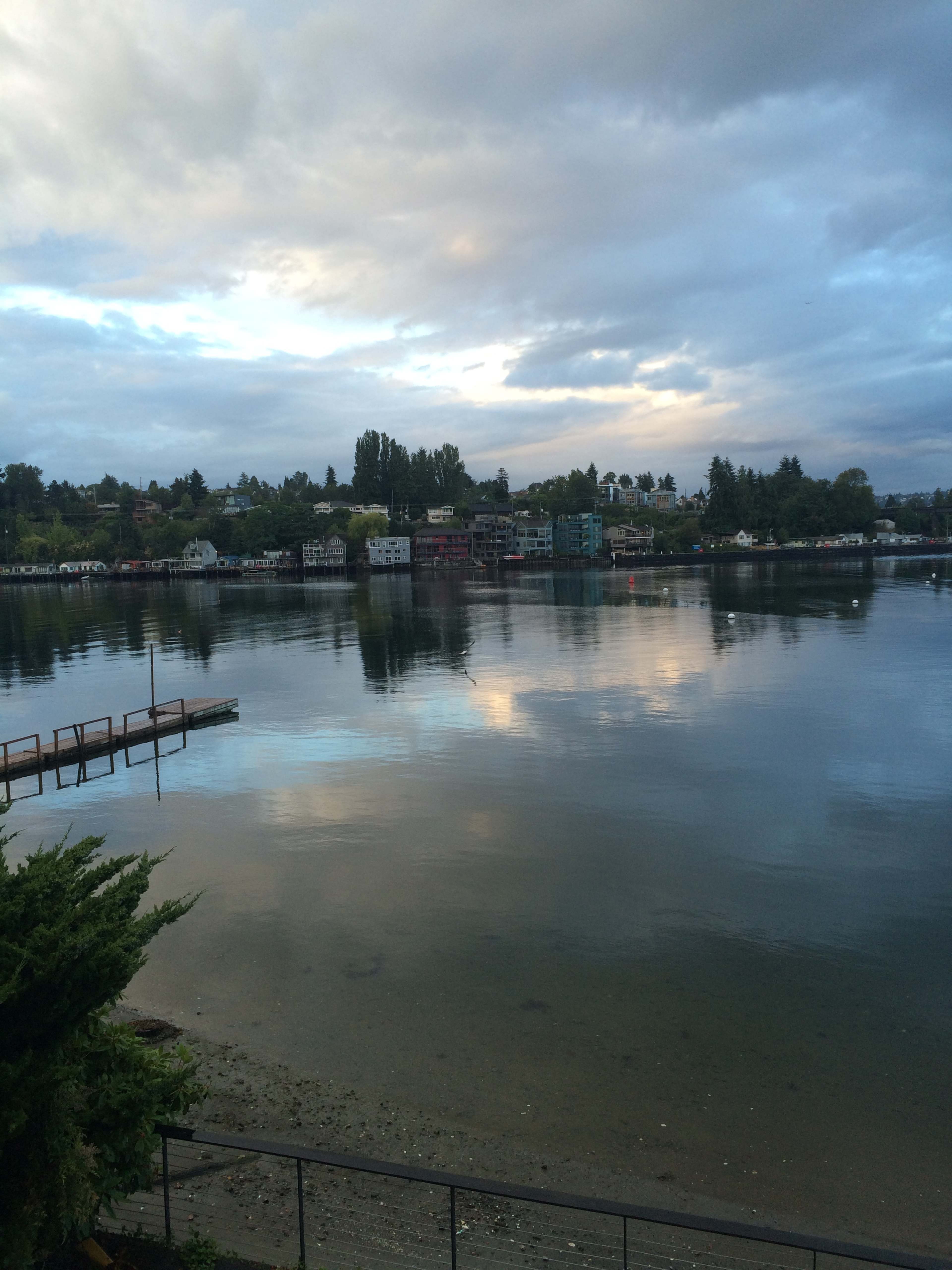 The image shows a calm body of water reflecting clouds and houses along the shoreline under a cloudy sky.