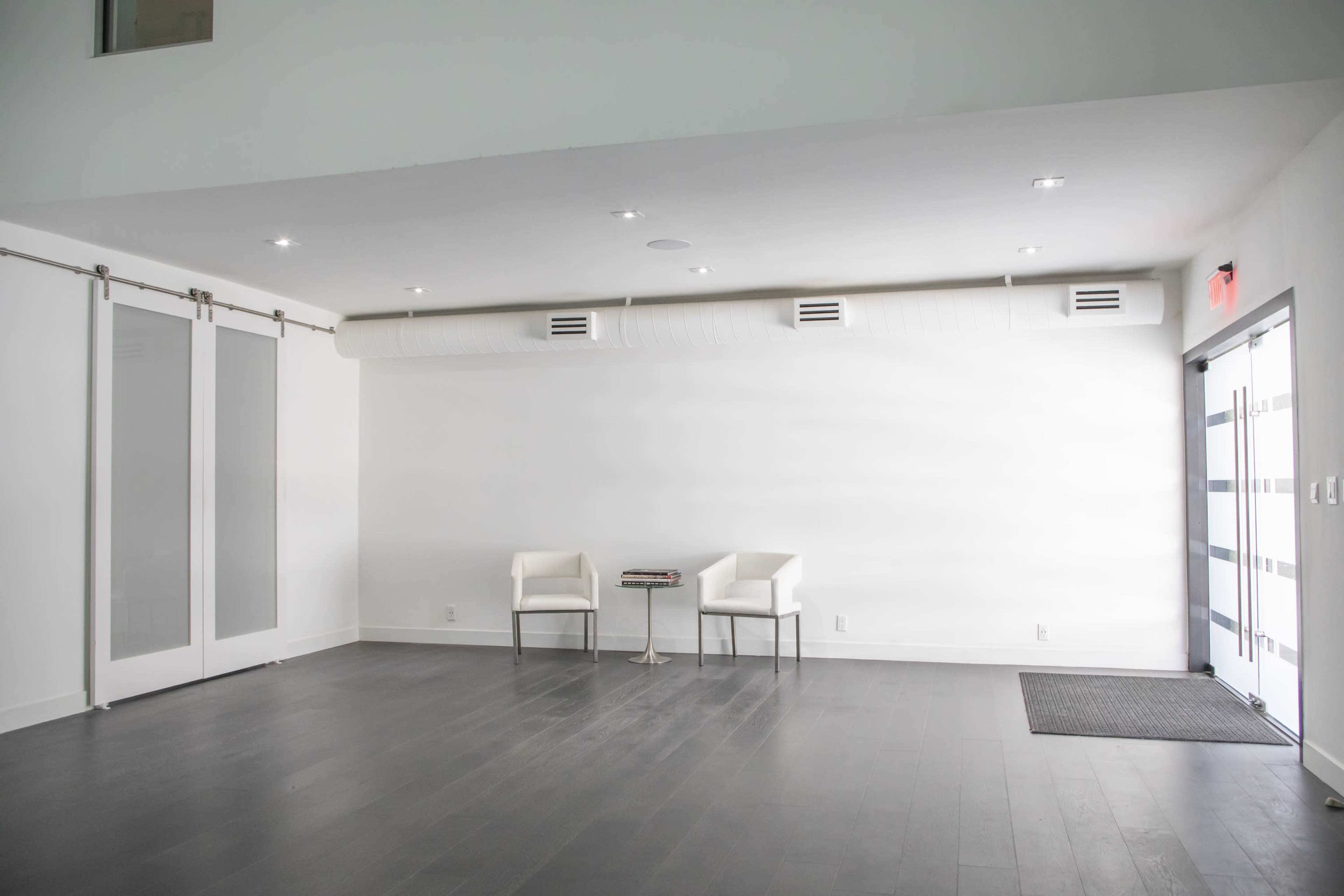 An empty interior with light gray walls, a dark wooden floor, two white chairs next to a small table, and a large glass door.
