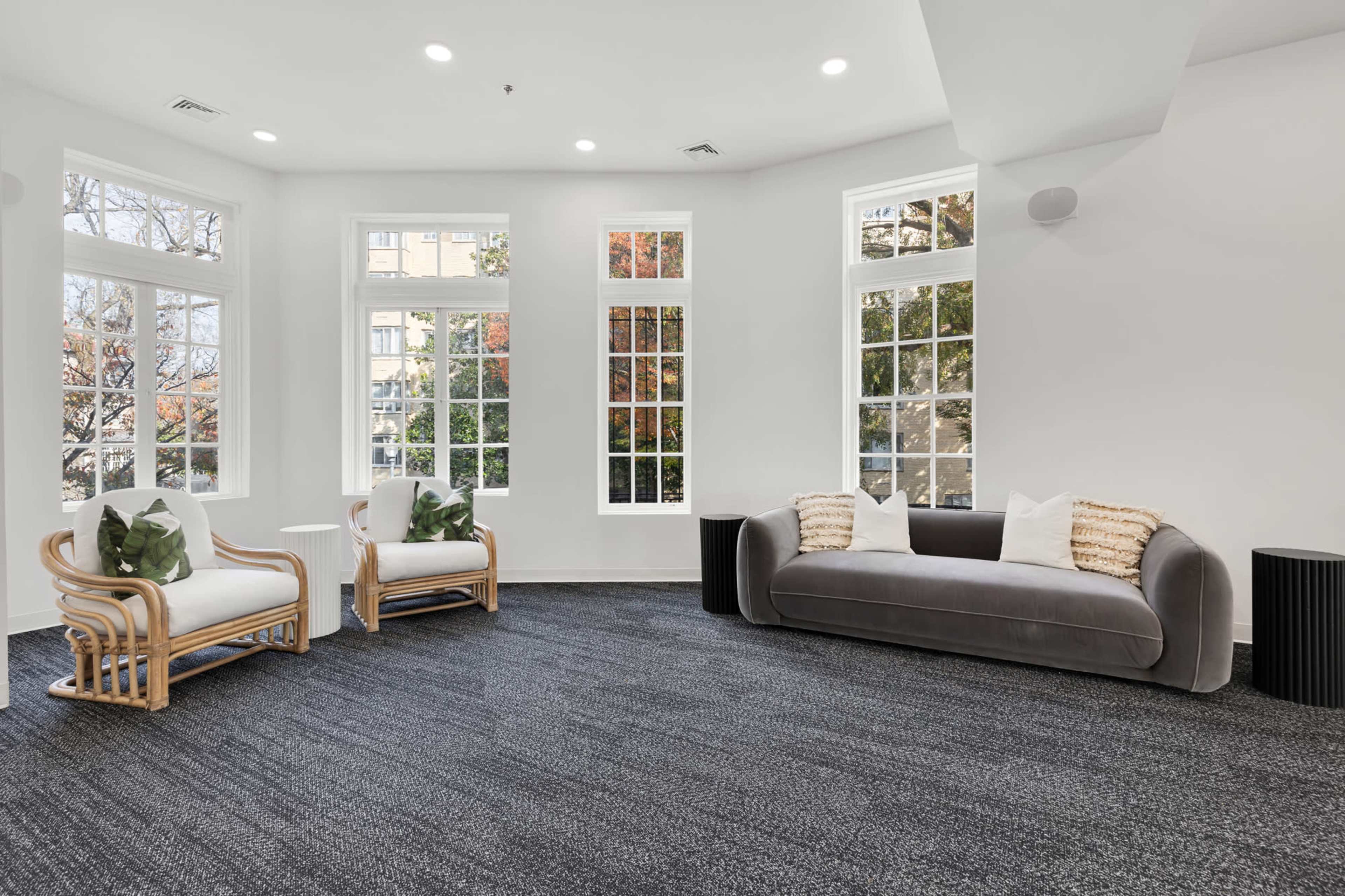 A bright room features two rattan chairs and a plush gray sofa, surrounded by large windows that offer a view of trees.