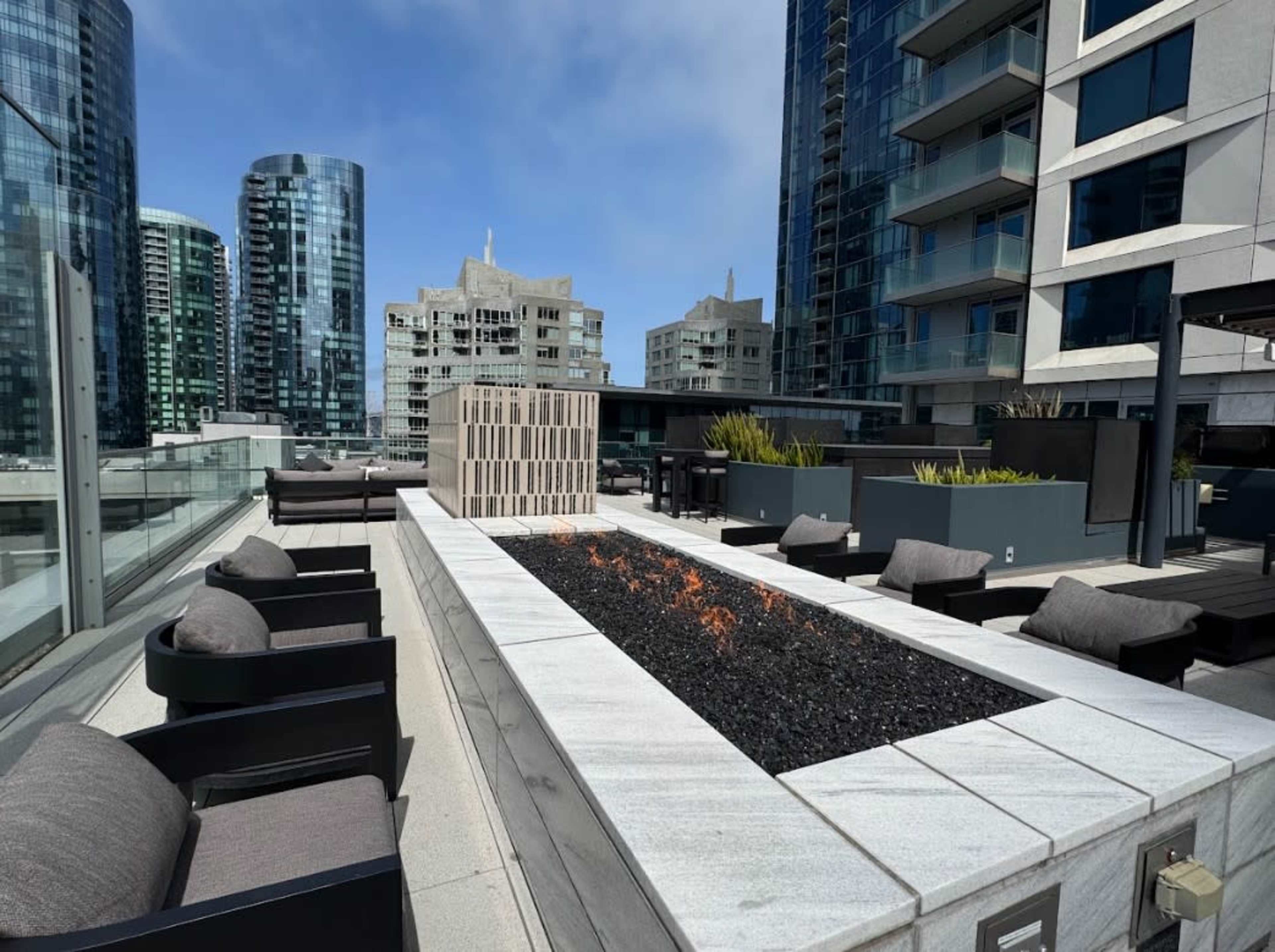 The image shows a rooftop seating area featuring a modern fire pit surrounded by comfortable chairs and city skyscrapers in the background.