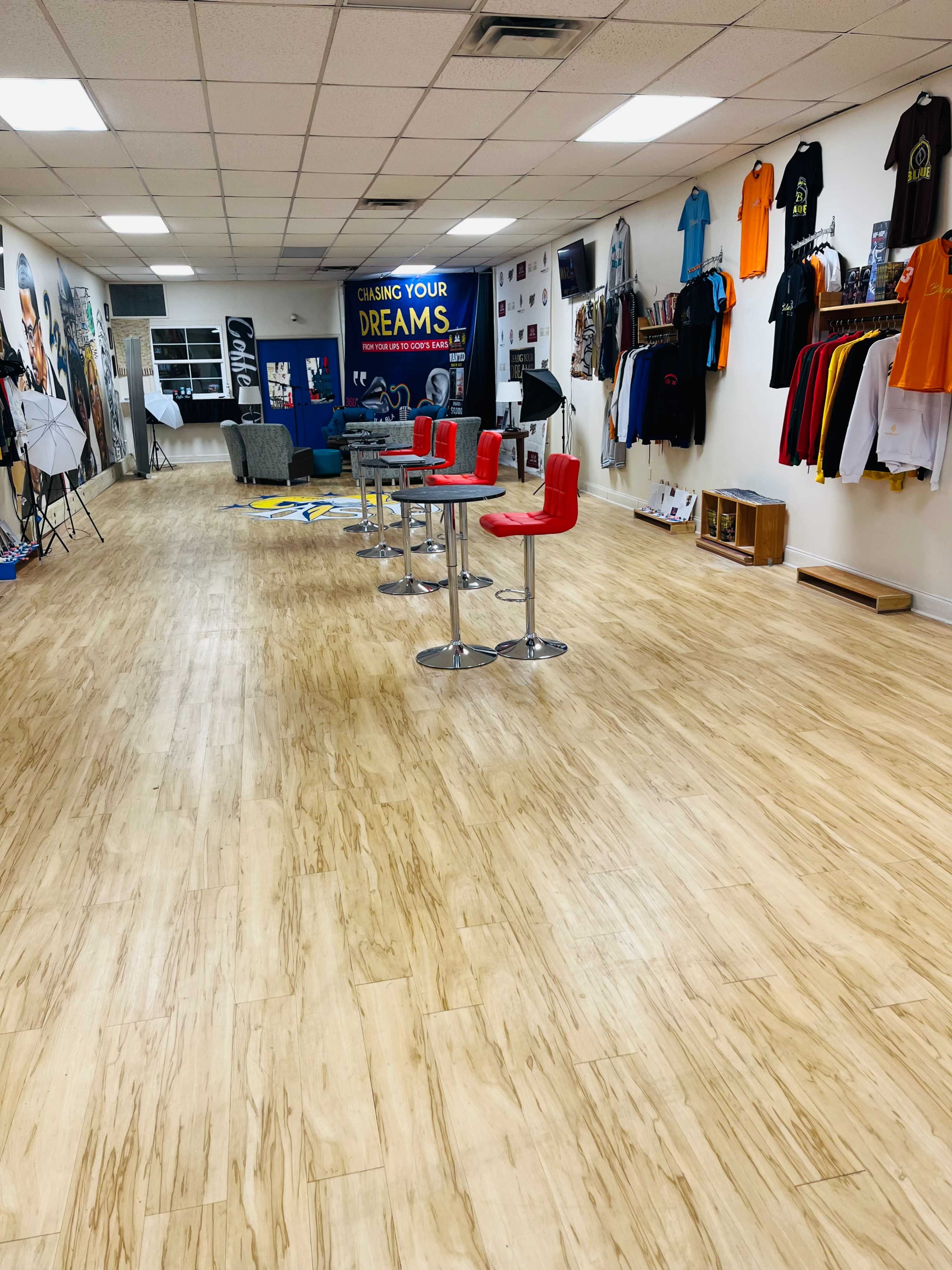 The image shows a spacious room with wooden flooring, featuring several red bar stools arranged around small tables and walls adorned with various T-shirts and motivational posters.