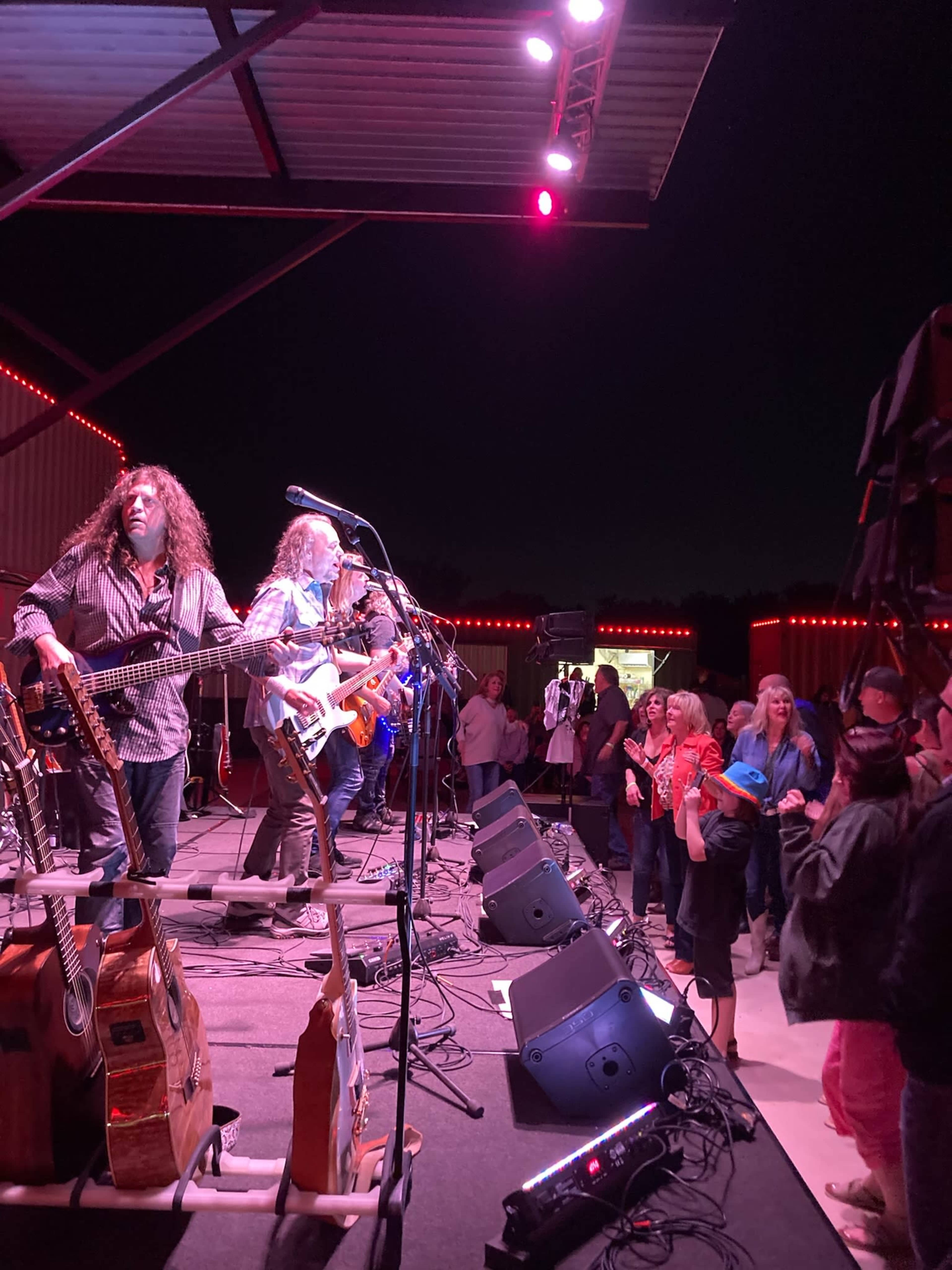A band performs on stage with colorful lights as an audience enjoys the live music in an outdoor setting at night.