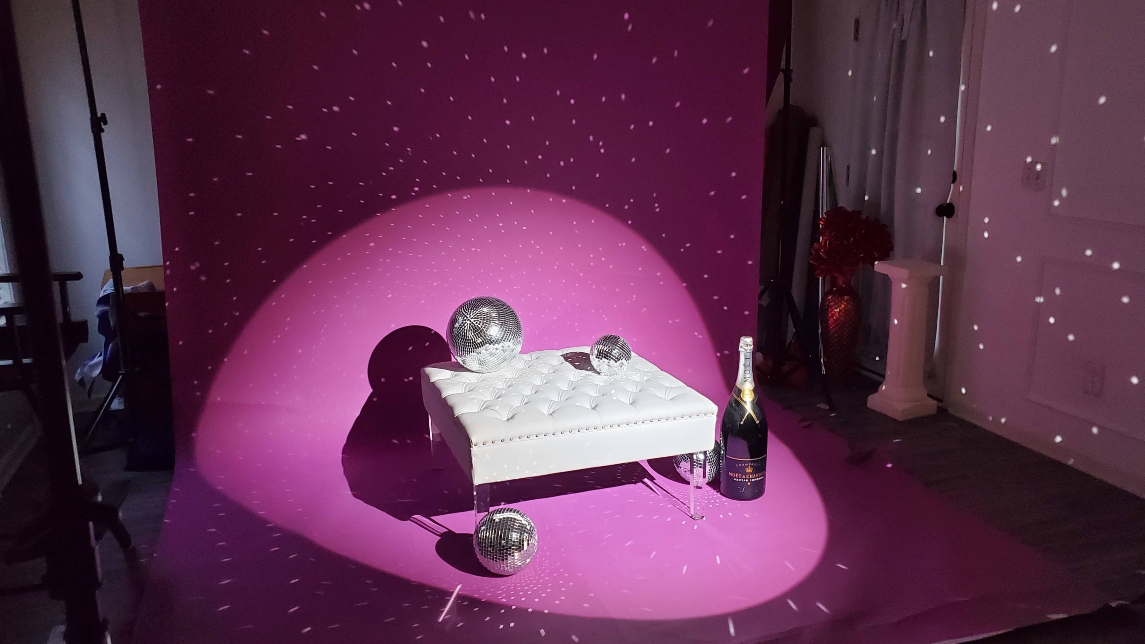 The image features a white tufted ottoman with two disco balls and a bottle of champagne, all set against a vibrant pink backdrop illuminated by scattered light reflections.