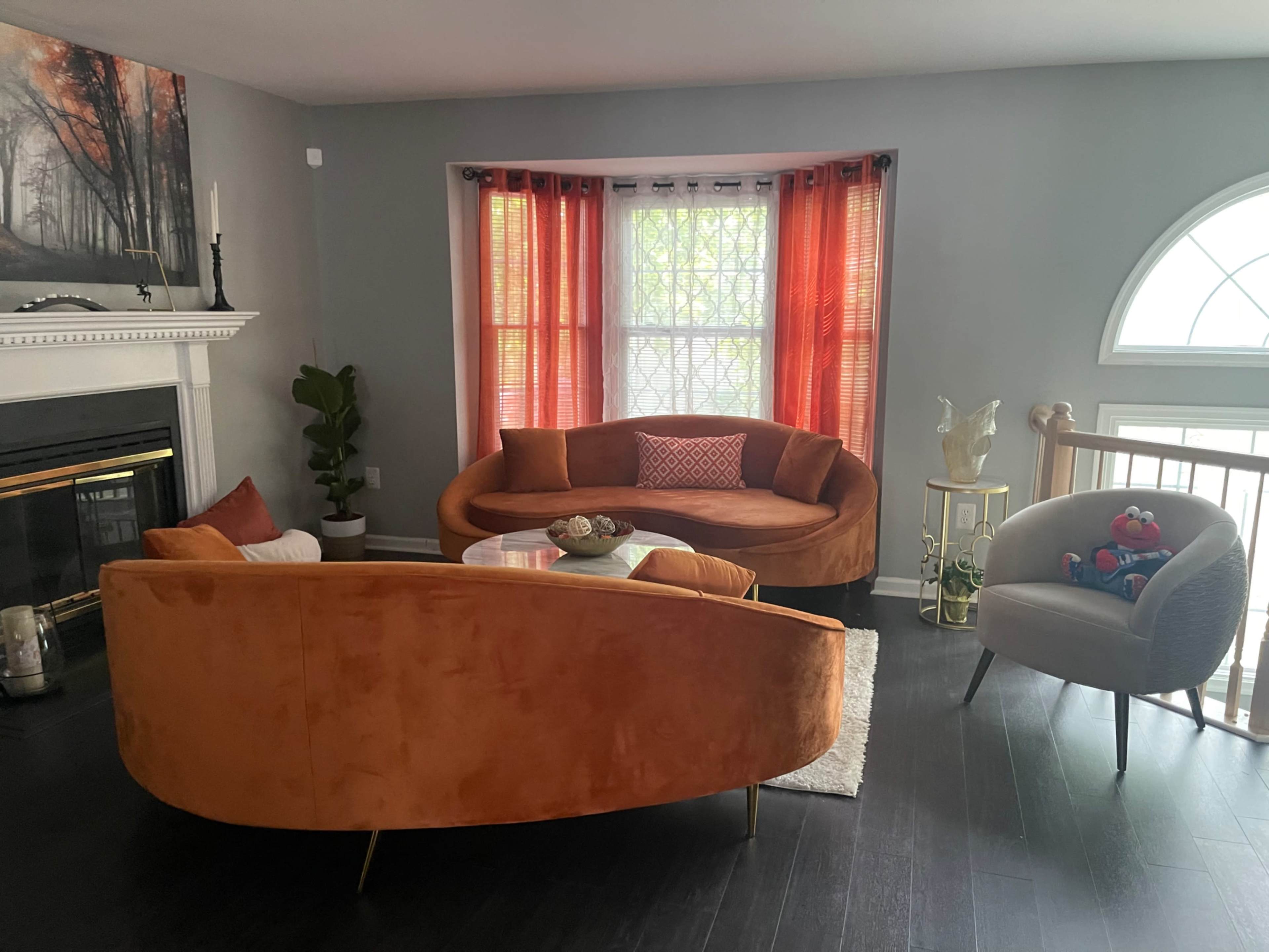 A modern living room with a circular orange sofa, a gray armchair, a small glass coffee table, and large windows draped with orange curtains.