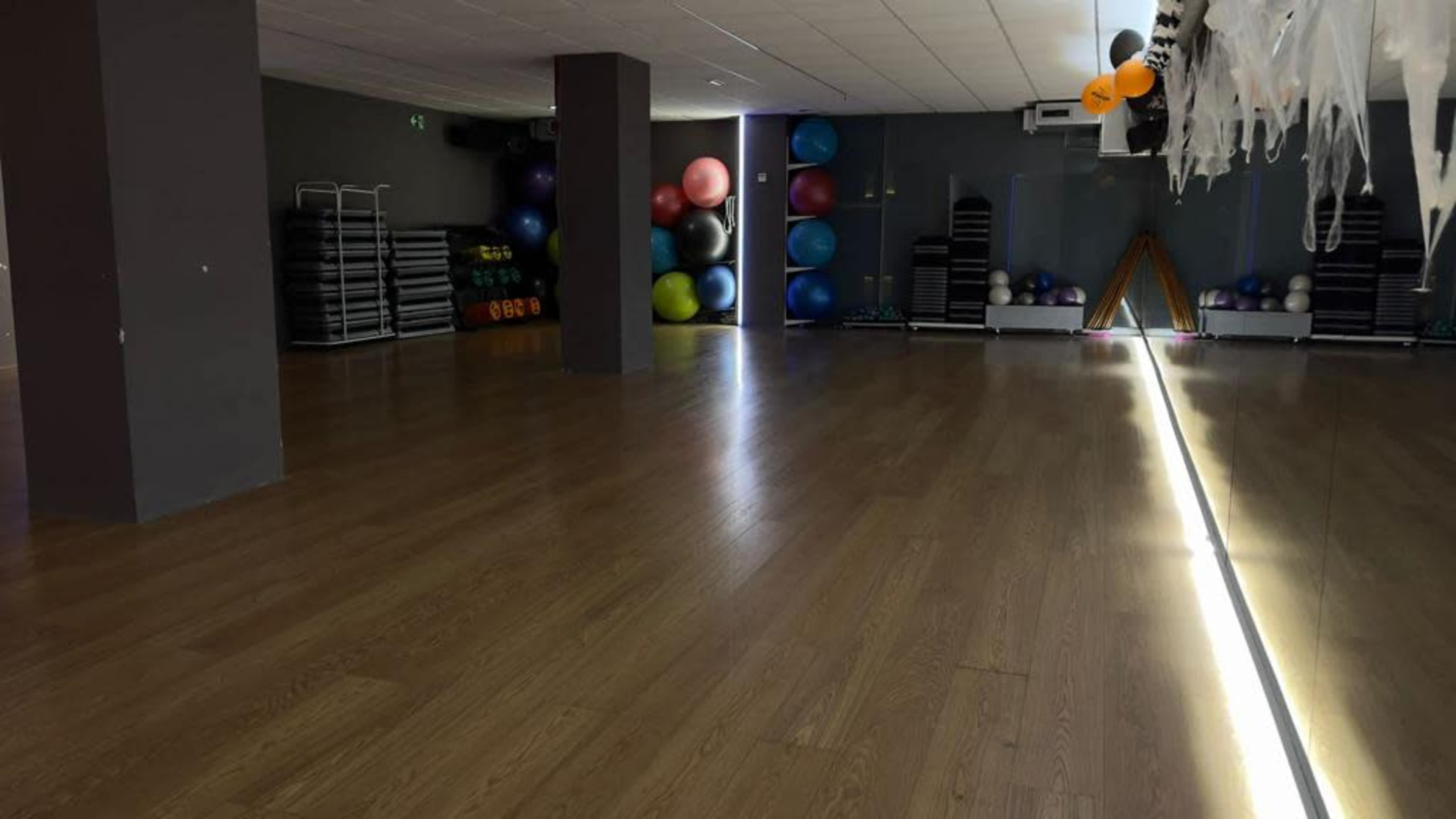 The image shows an empty gym studio with wooden flooring and fitness equipment, including colorful exercise balls and weights, arranged against the walls.