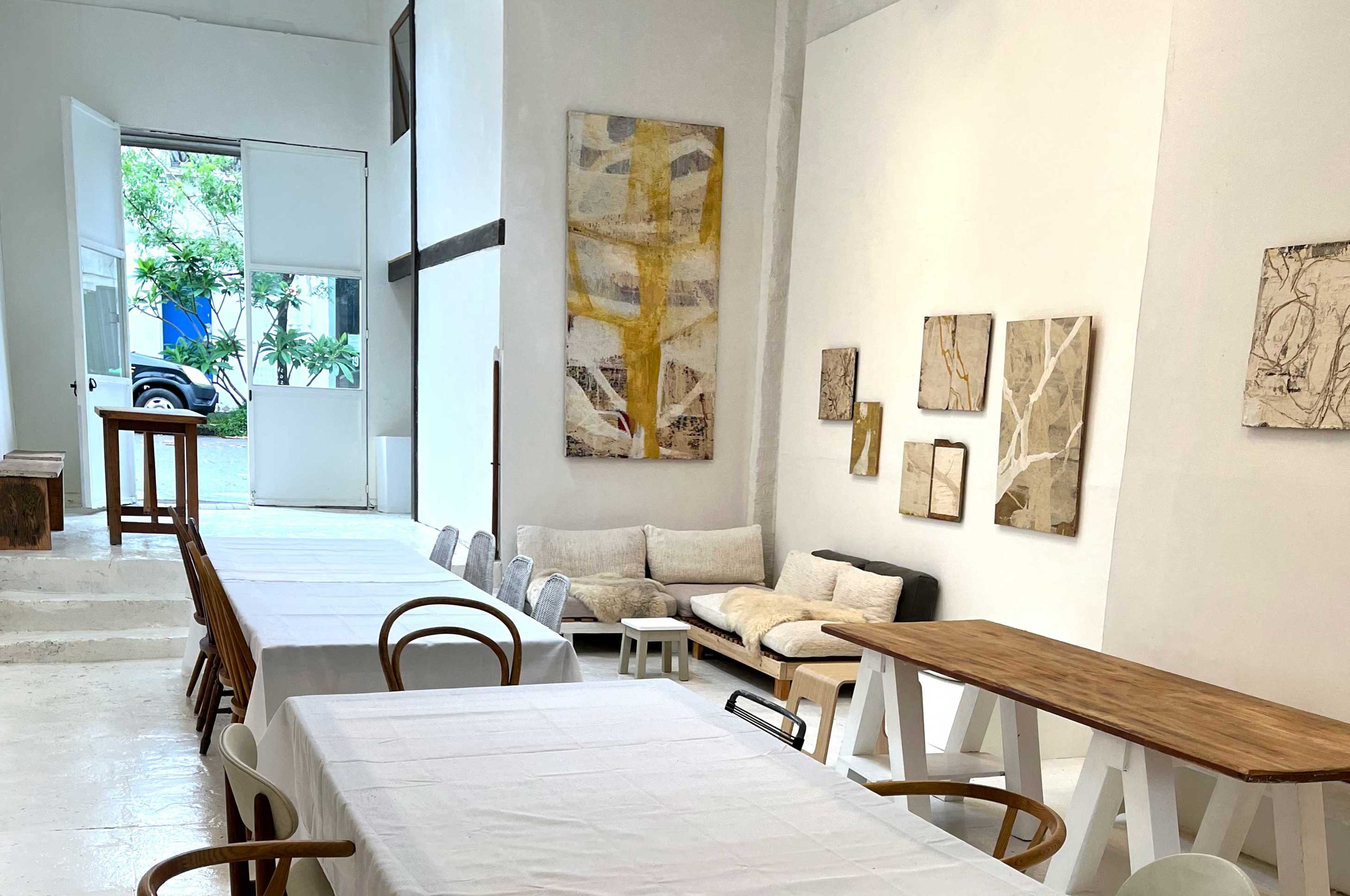 The image shows a spacious, minimalist interior with tables and chairs, a sofa, and artworks on the walls, leading to an open courtyard.