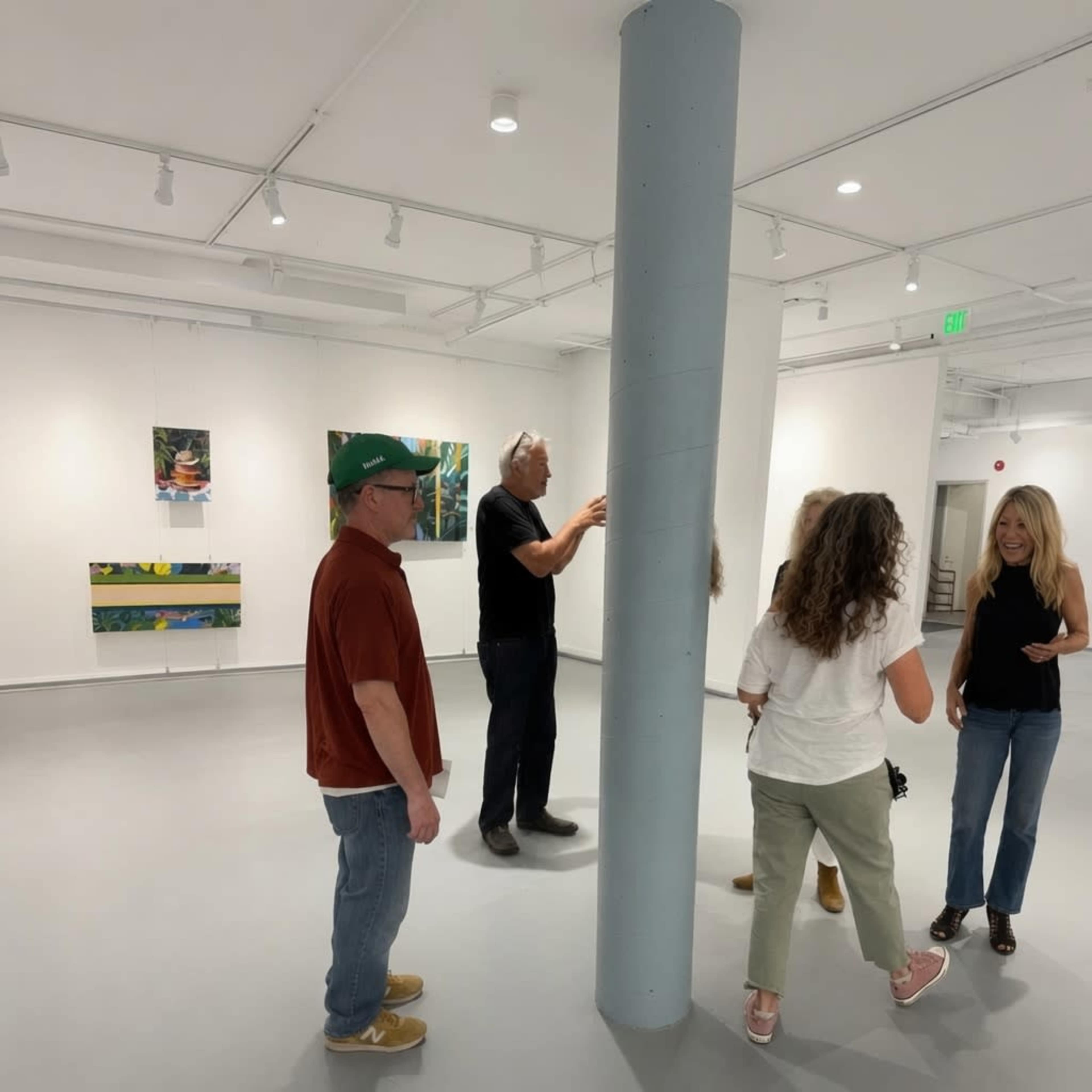A group of five people interacts in an art gallery, with several paintings displayed on the white walls.