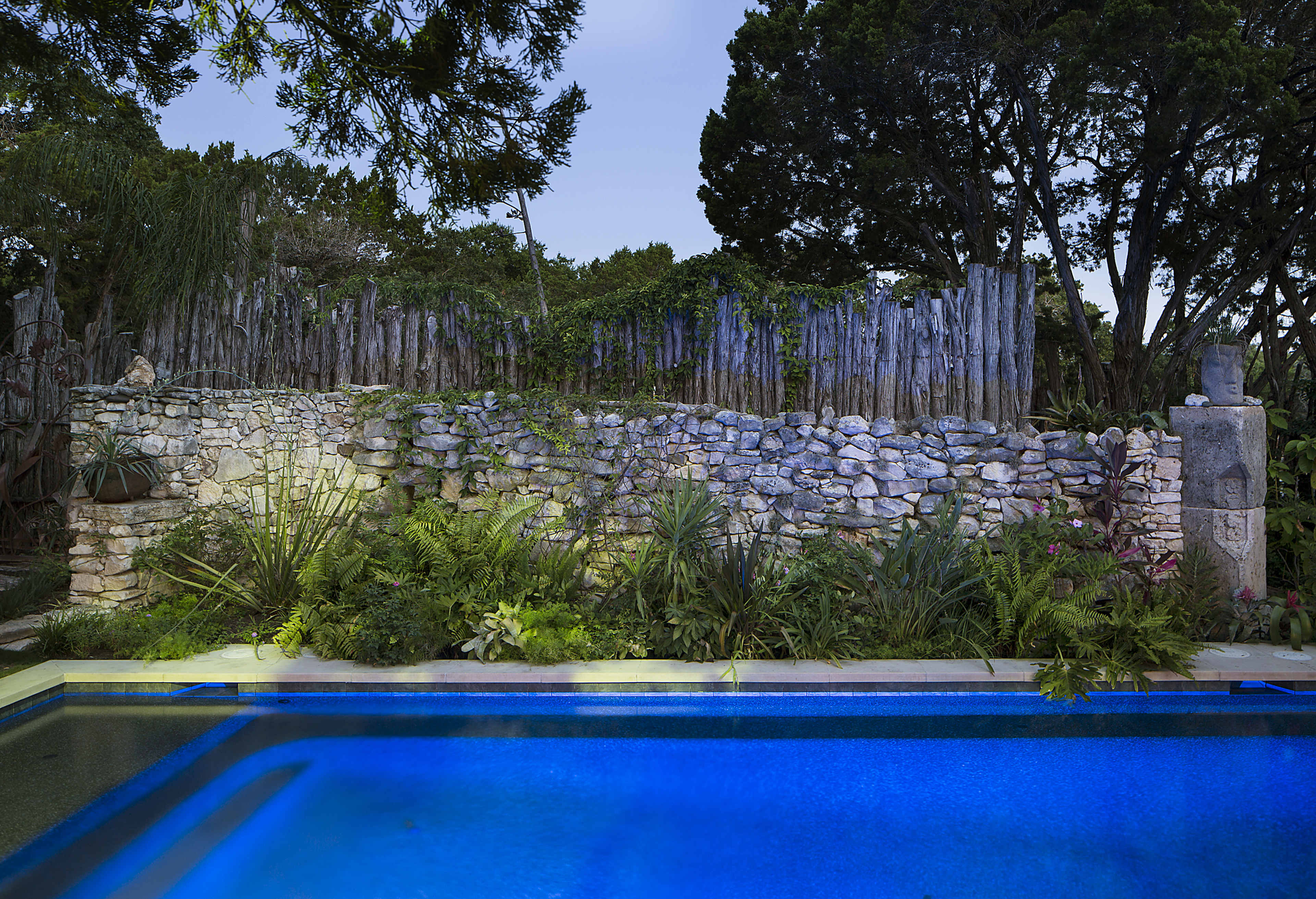 A blue swimming pool in the foreground, bordered by lush greenery and a stone wall with a wooden fence in the background under a twilight sky.
