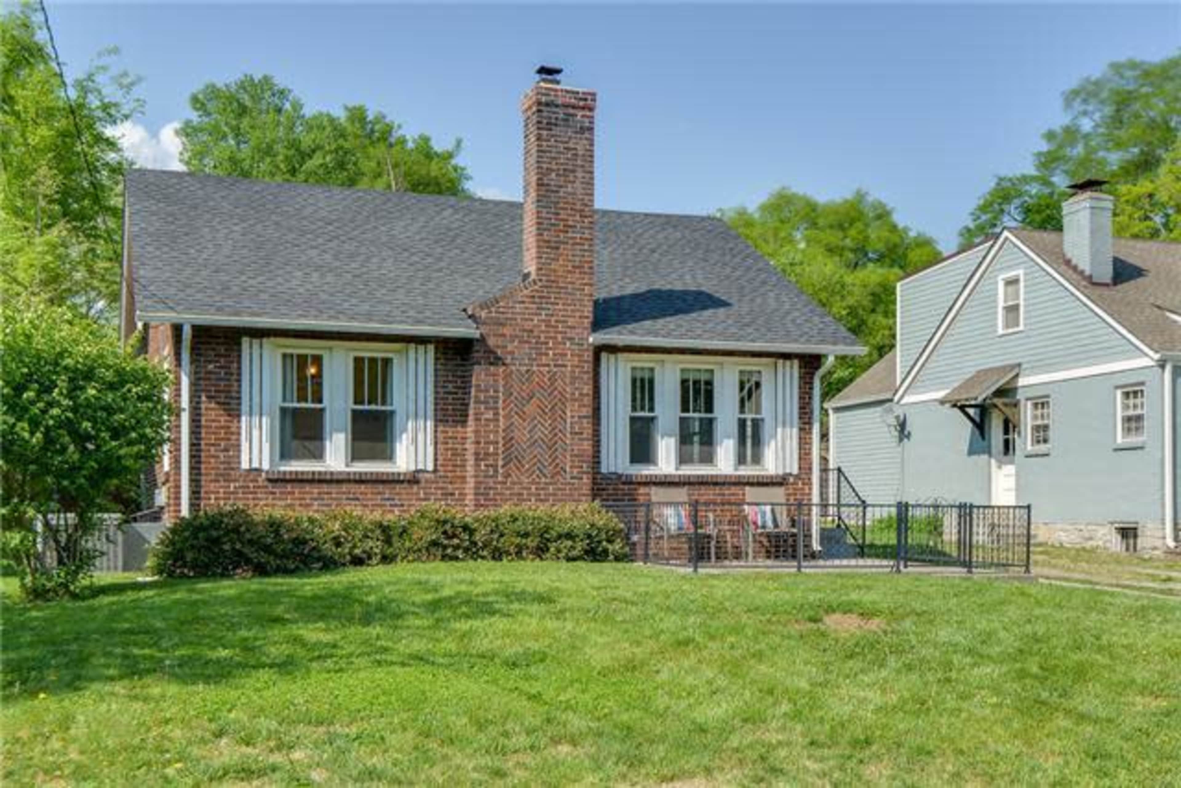 A brick house with a chimney and front porch is surrounded by a green lawn and nearby homes.