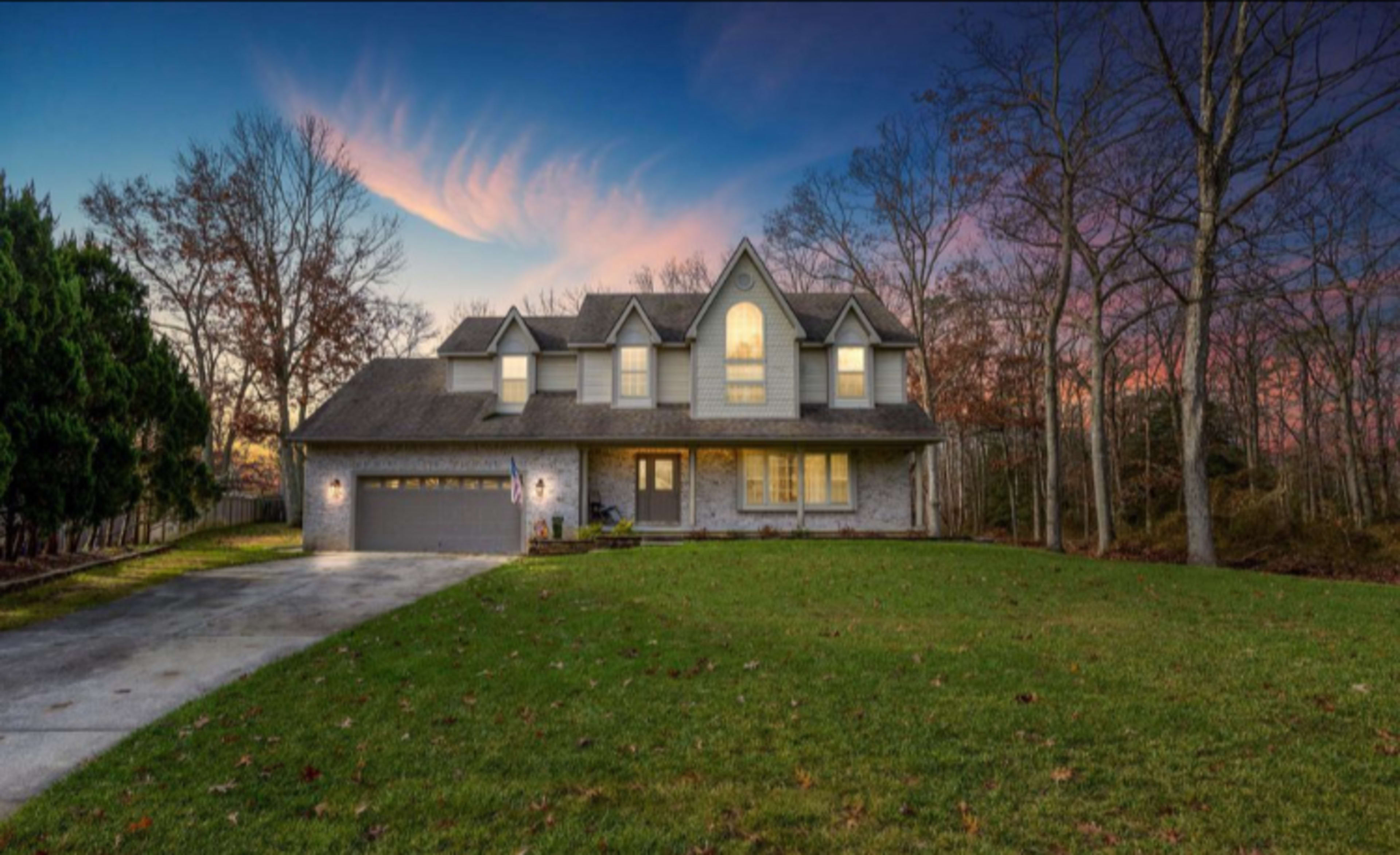 A large two-story brick house with a landscaped front yard sits under a colorful sunset sky amidst trees.