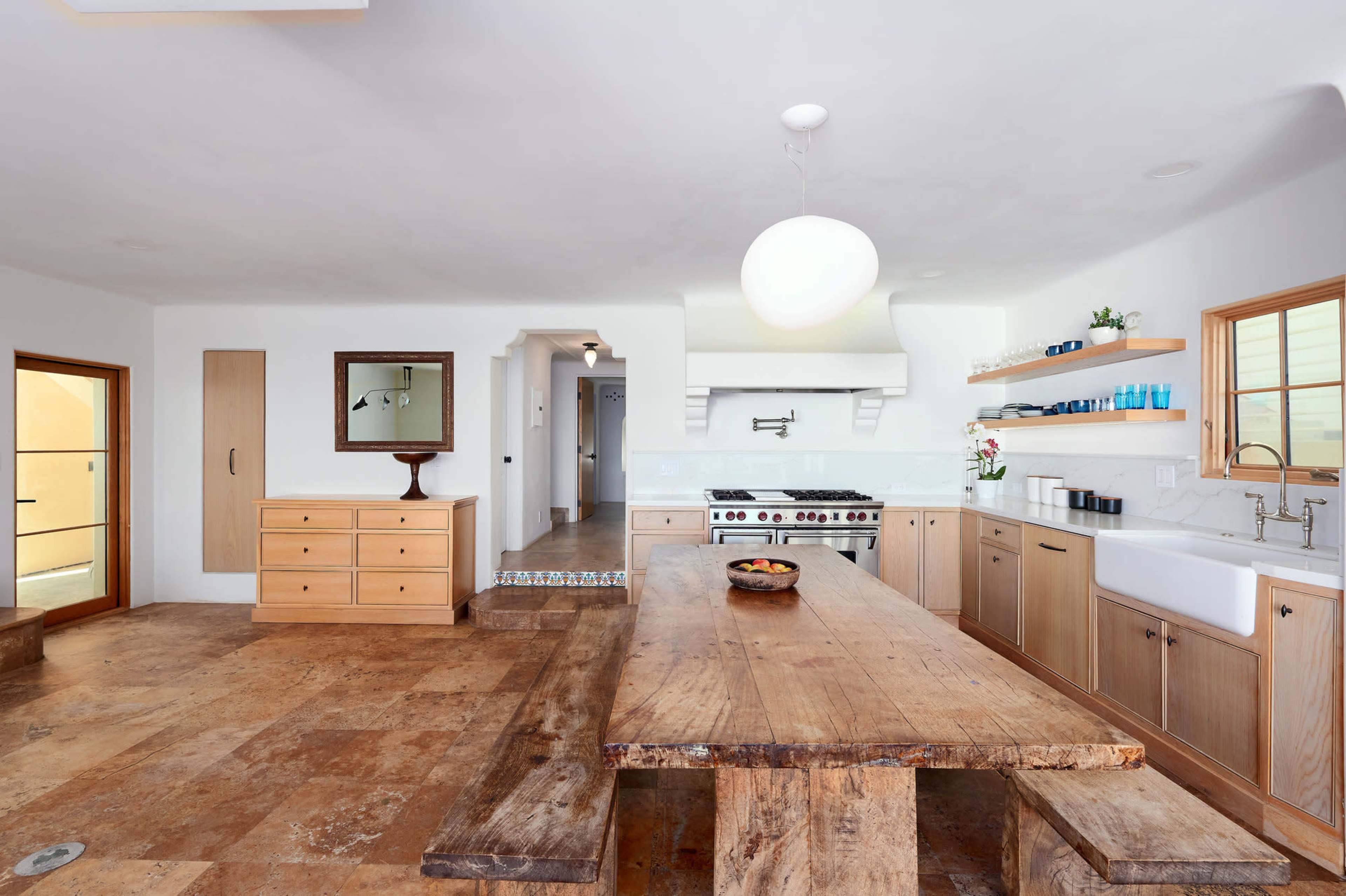 The kitchen features a large wooden dining table, a farmhouse sink, and modern appliances against a backdrop of light-colored walls and cabinets.