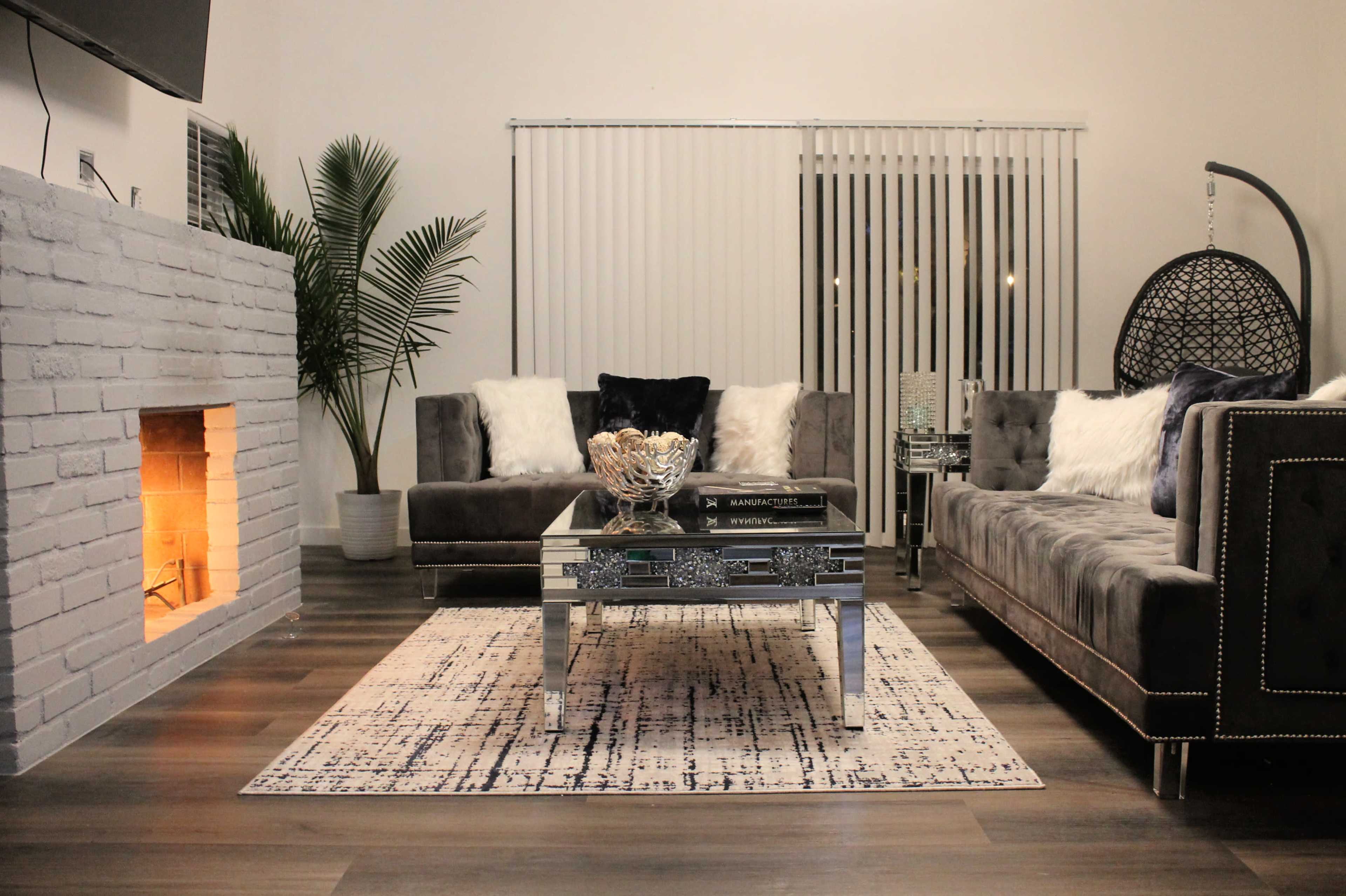 A modern living room features a gray sectional sofa, a mirrored coffee table, and a cozy fireplace, with a plant in the corner and large windows adorned with vertical blinds.