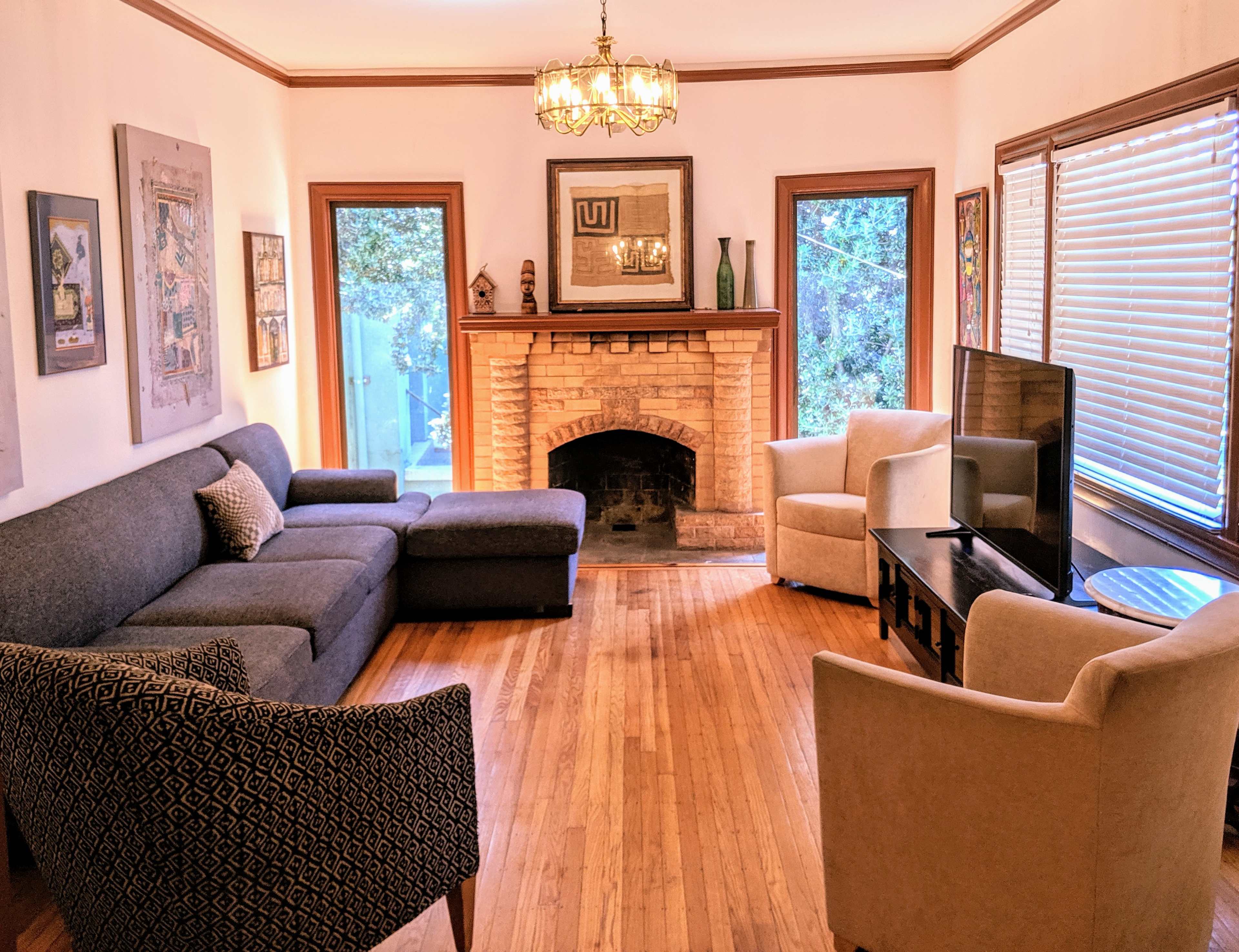 A living room with a gray sectional sofa, two light-colored armchairs, a television, and a brick fireplace, featuring large windows that allow natural light.