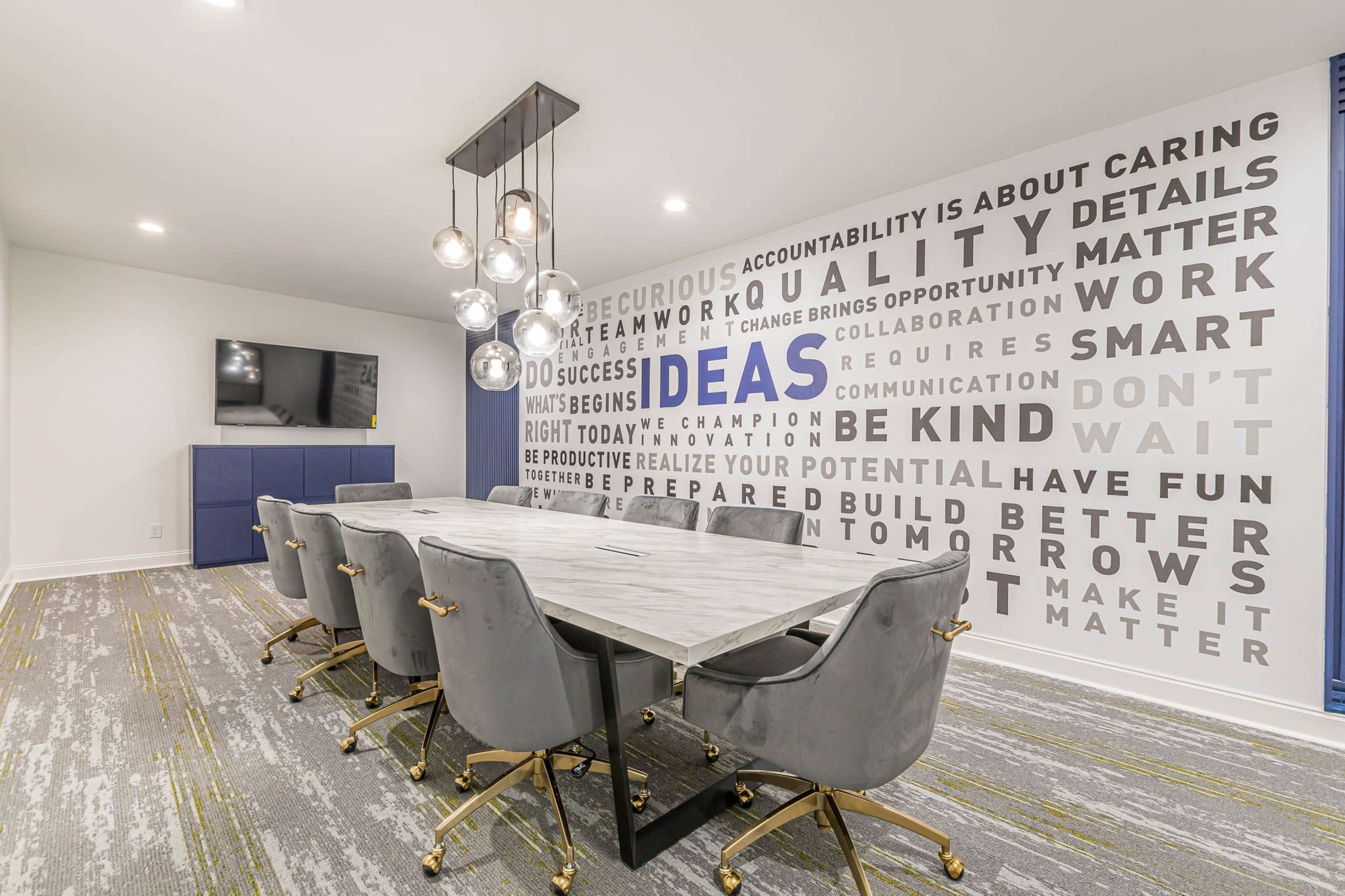A modern meeting room features a large table with gray chairs, a wall adorned with motivational words, and a television setup in the corner.