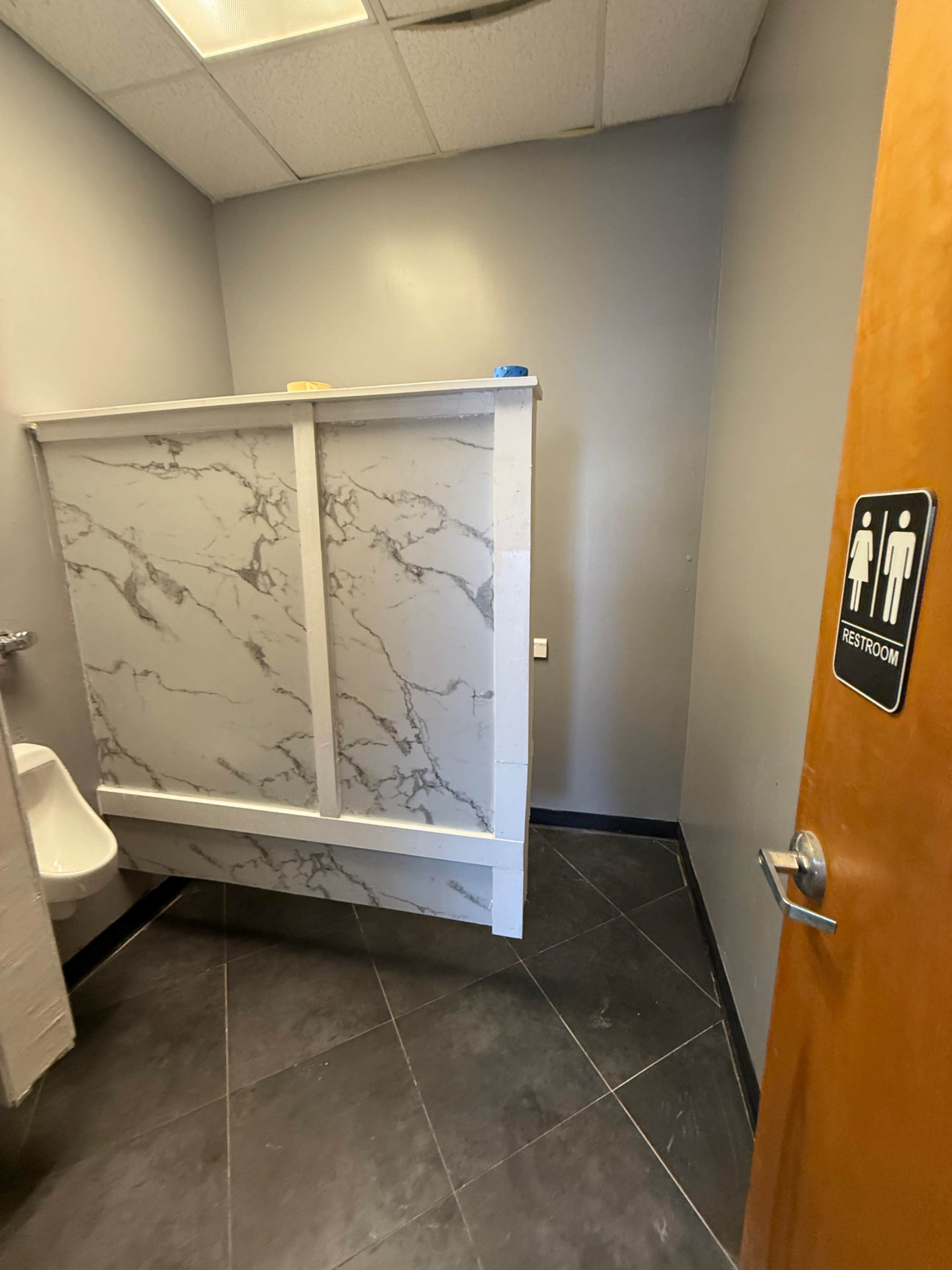 The image shows a bathroom interior with a partition made of marble-patterned material, a urinal on the left, and a door labeled "RESTROOM" on the right.