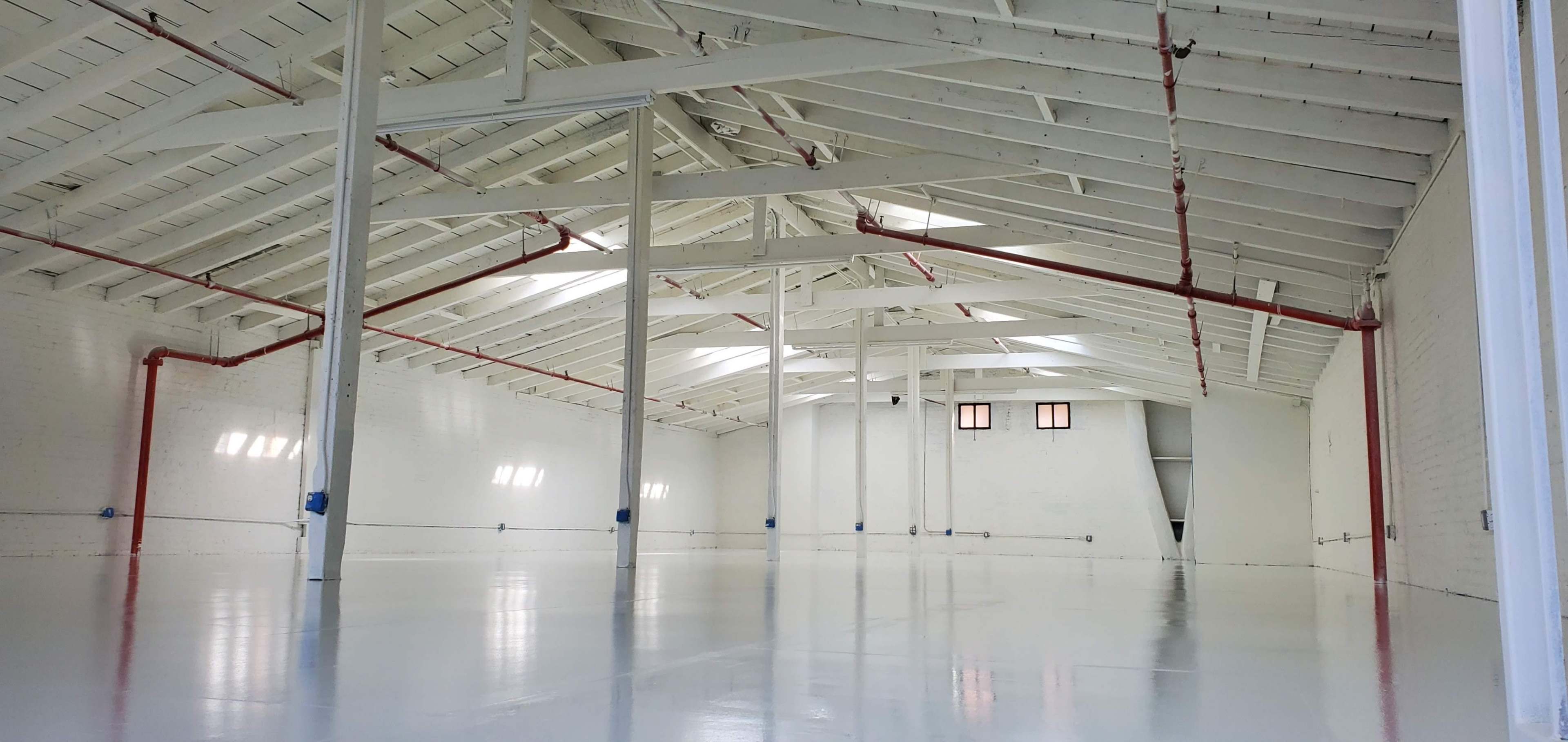 A large, empty interior space with a high, white ceiling, exposed beams, and a polished floor.