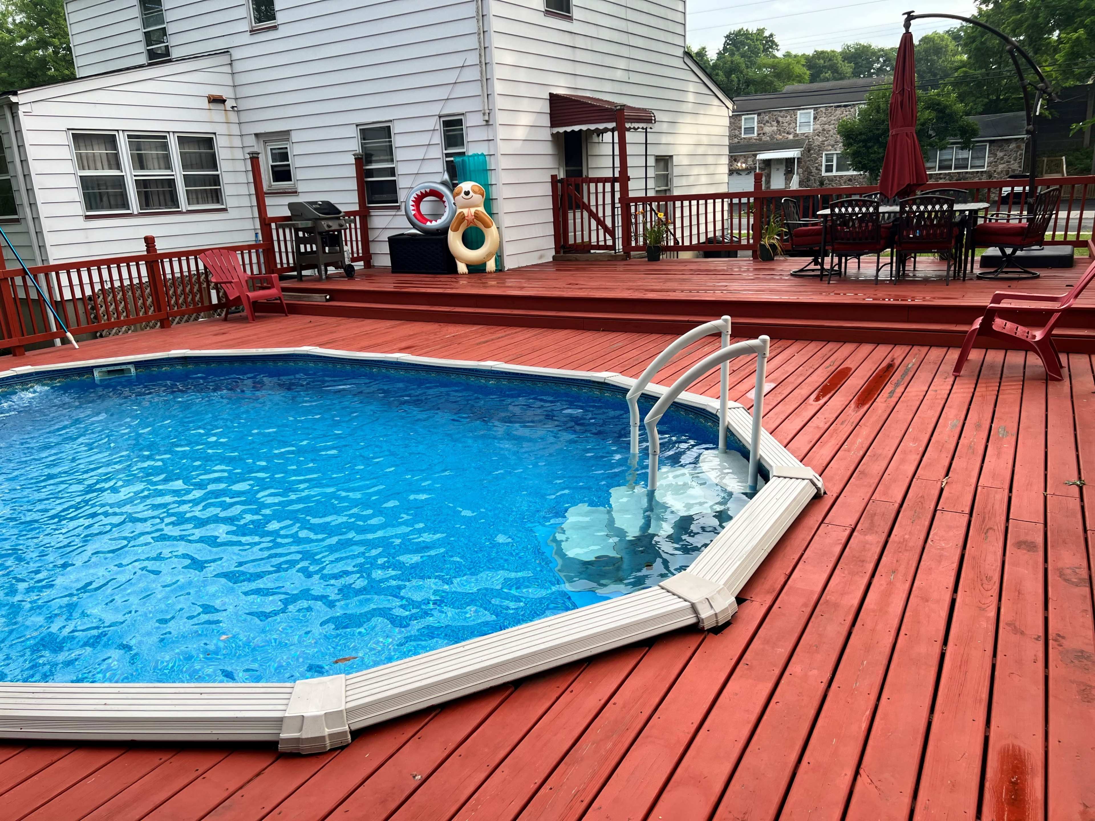 The image shows a backyard with a circular above-ground pool surrounded by a wooden deck, patio furniture, and a large red umbrella.