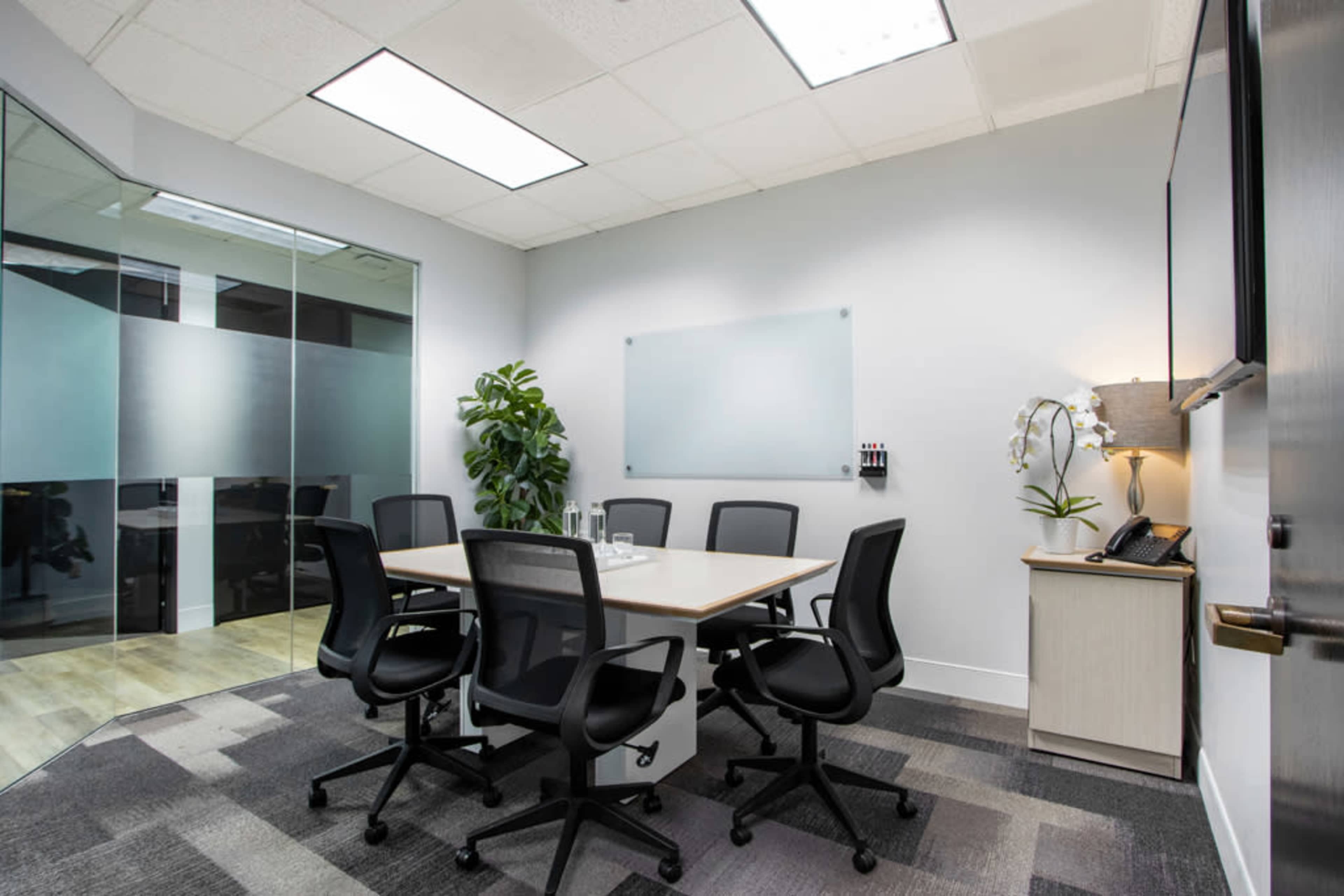 The image shows a modern conference room with a rectangular table surrounded by six black office chairs, a glass wall, and a wall-mounted phone.