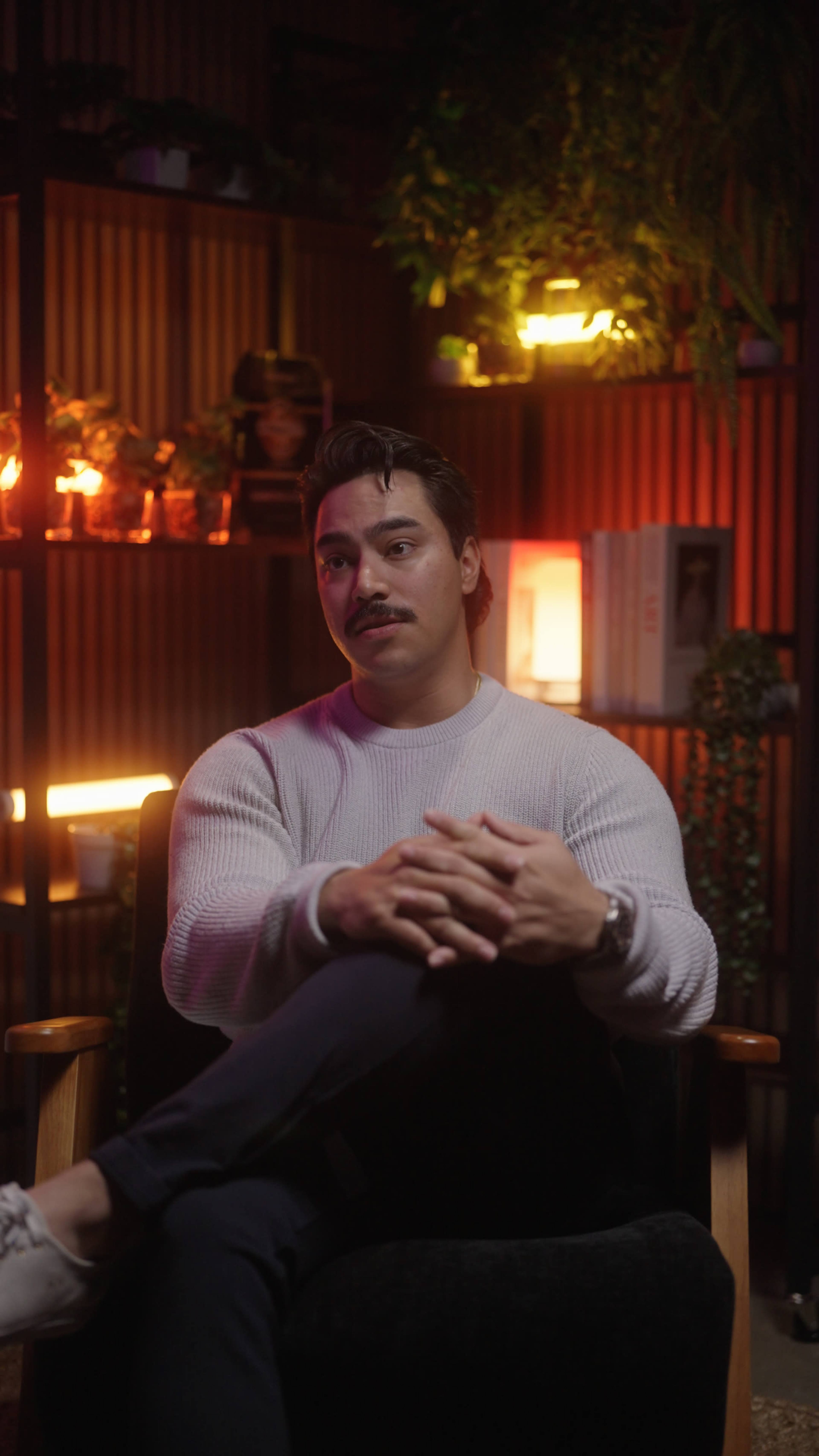 A man with a mustache sits in a chair with his hands clasped, surrounded by warm lighting and plants in the background.