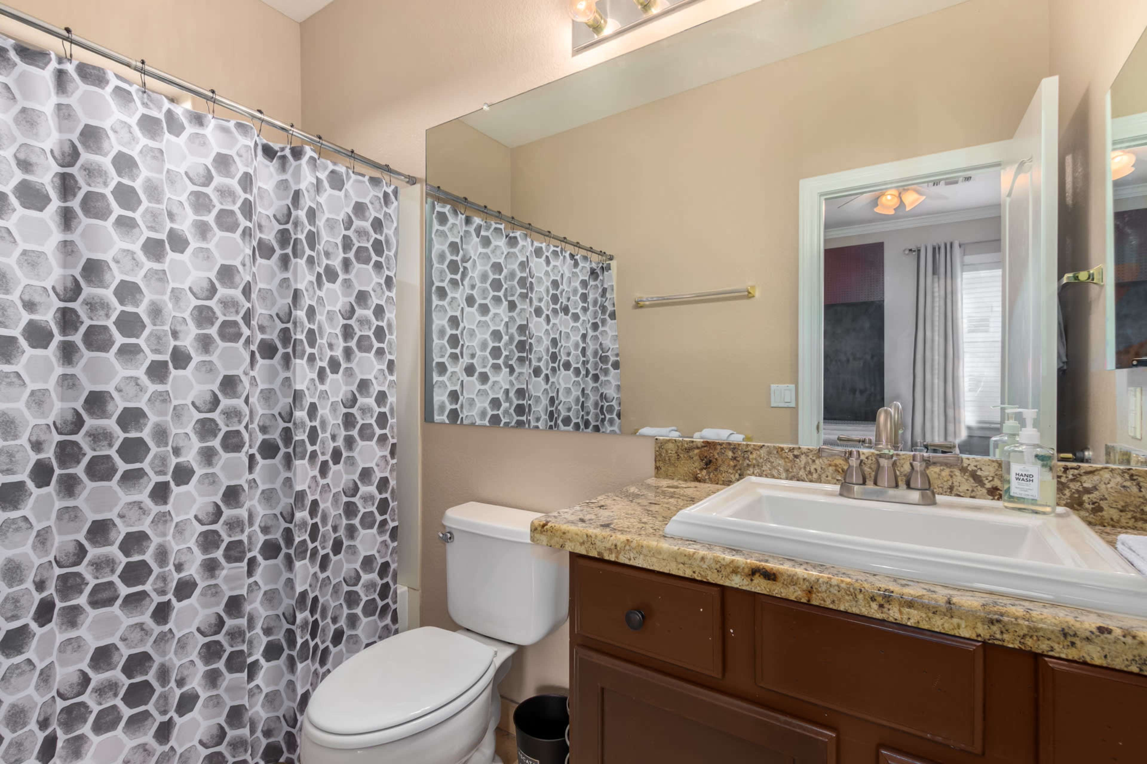 The image shows a bathroom featuring a granite countertop, a white sink, a toilet, and a shower curtain with a hexagonal pattern.