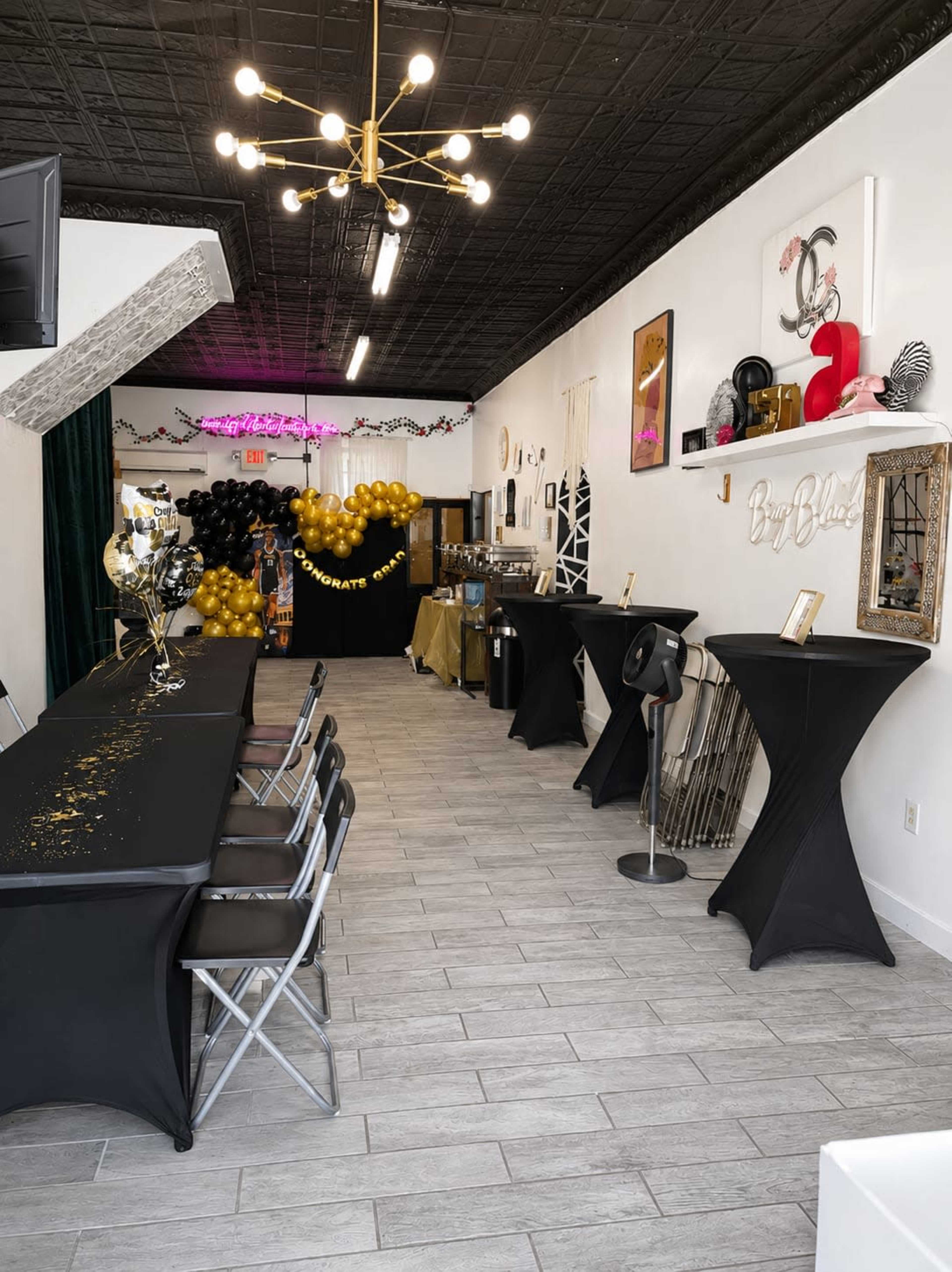 The image shows a decorated event space featuring black tables with gold accents, a photo backdrop with black and gold balloons, and festive lighting.