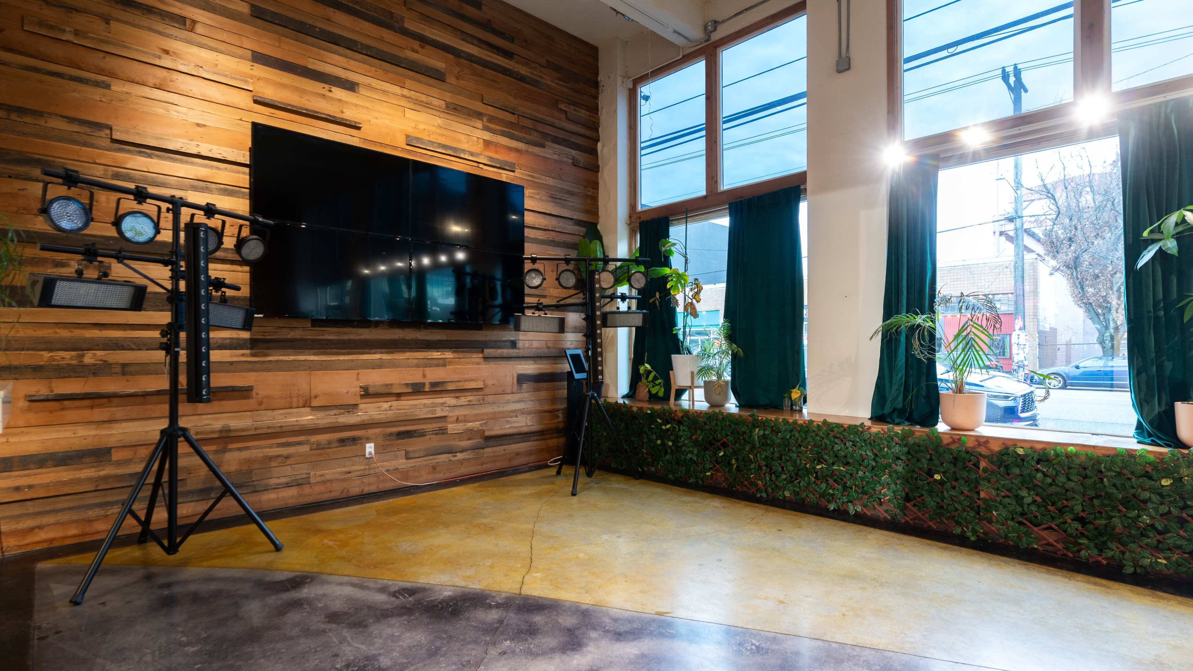 The image shows an interior space featuring a wooden wall, a large screen mounted on the wall, and lighting equipment on a stand, with windows overlooking a street outside.
