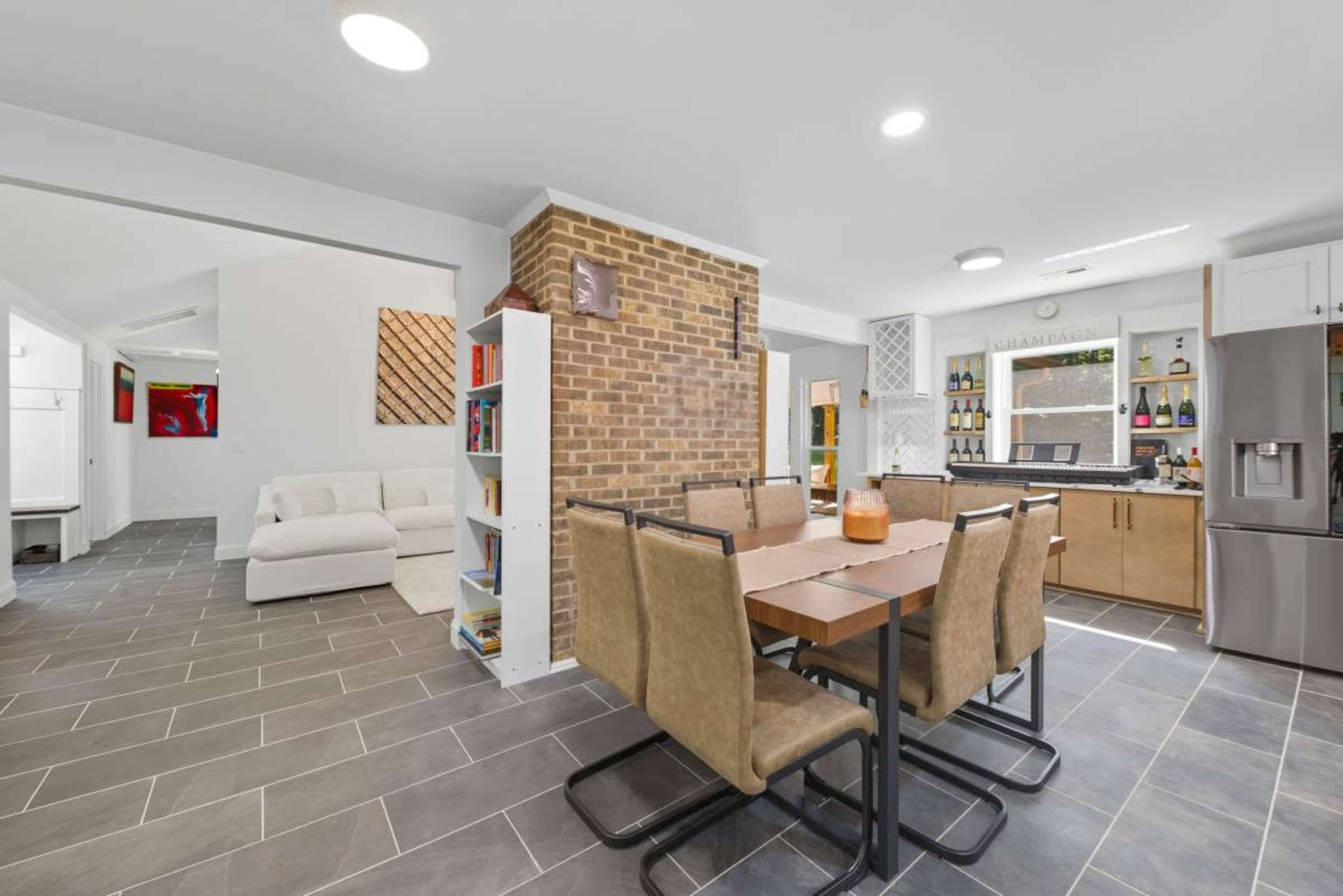 The image shows a modern kitchen and dining area with a wooden dining table surrounded by upholstered chairs, adjacent to a living space with a white sofa and bookshelves.