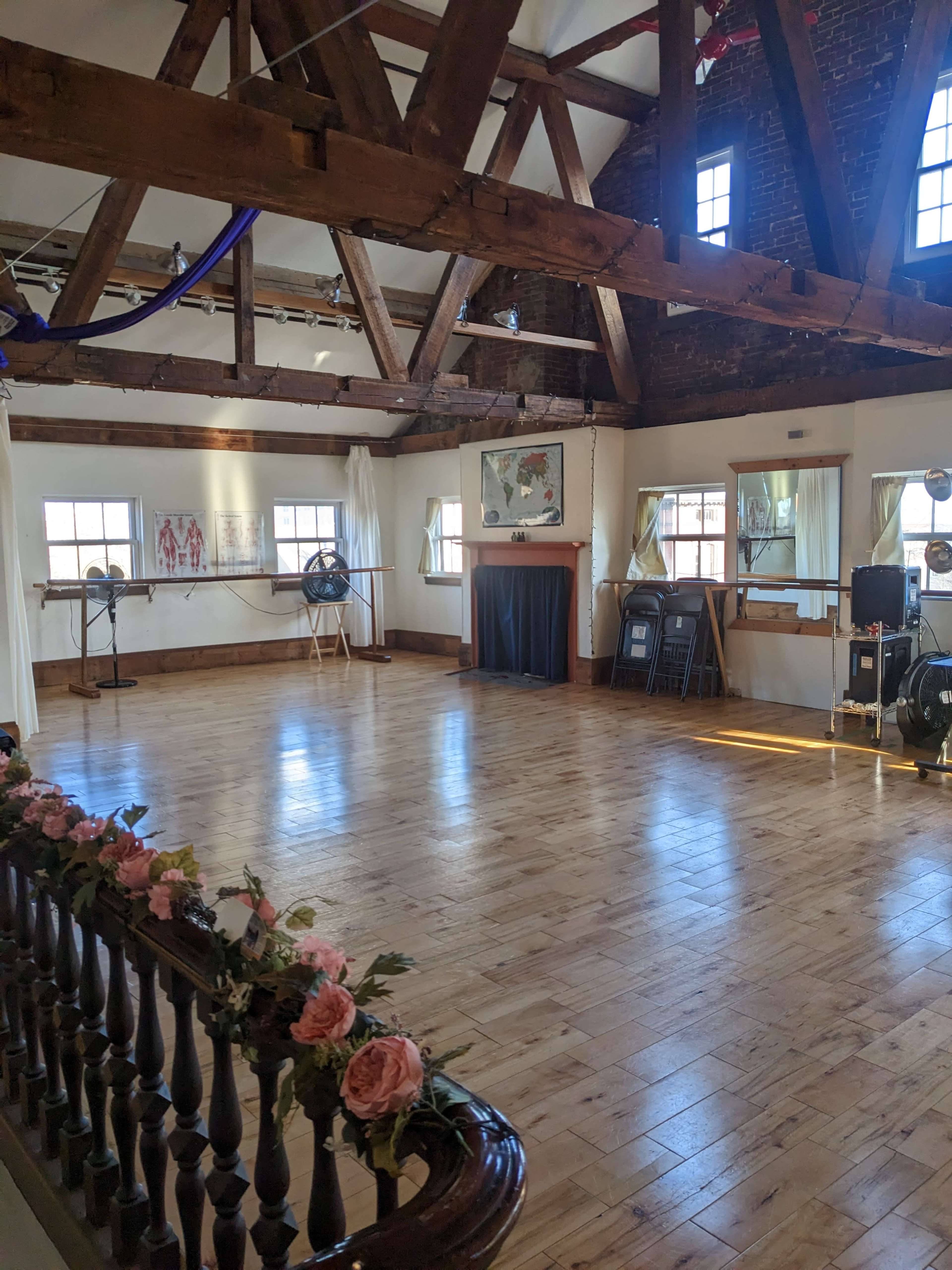 An empty dance studio with wooden floors, exposed beams, and large windows letting in natural light.
