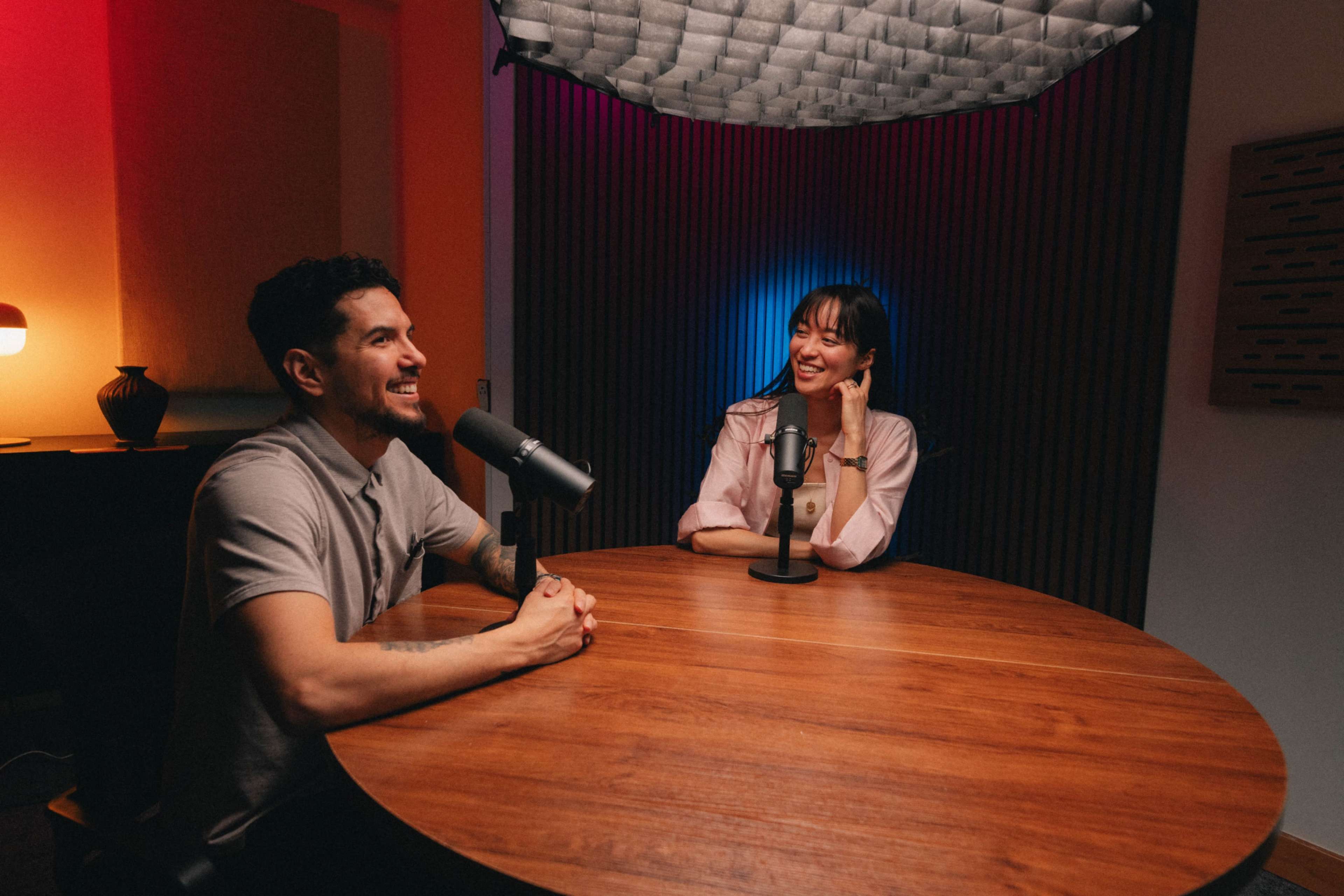 A man and a woman sit at a round wooden table with microphones in front of them, engaged in conversation in a well-lit studio environment.