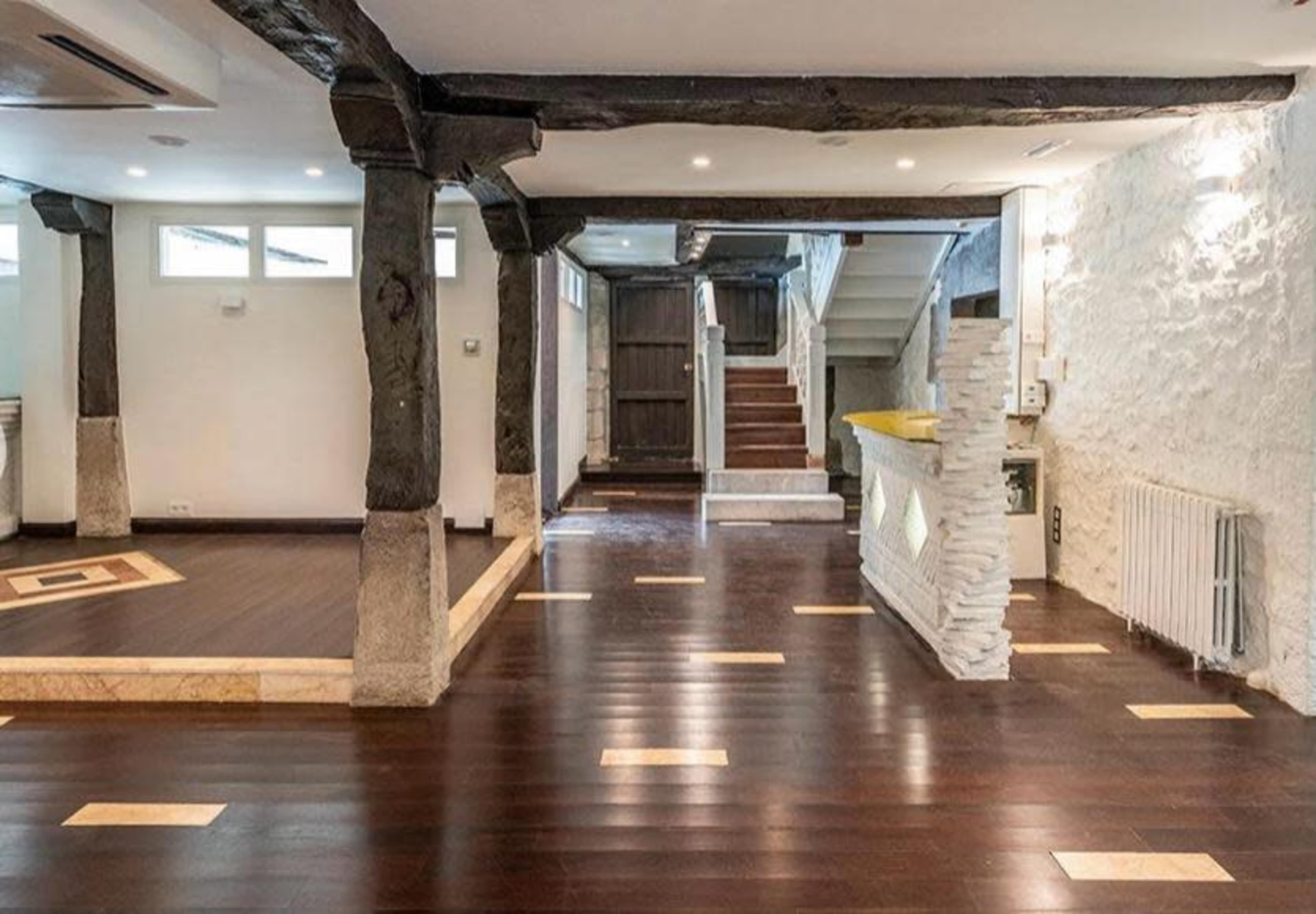 The image shows a spacious interior of a modern room featuring hardwood flooring, exposed wooden beams, and a staircase in the background.